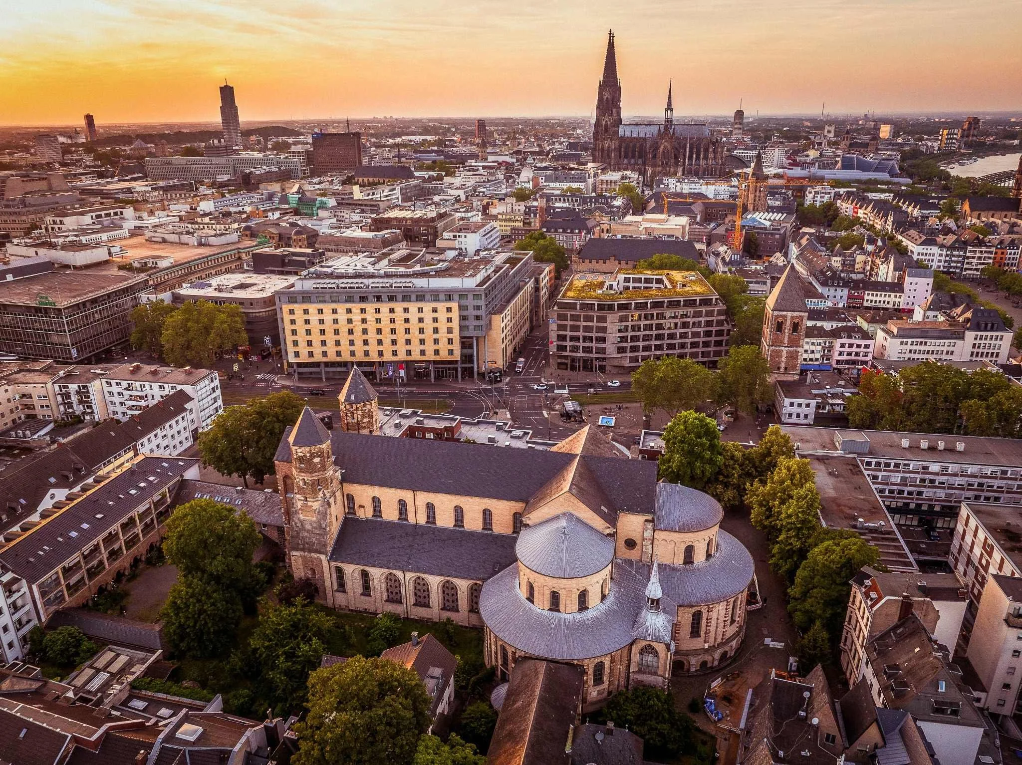 Luftaufnahme des Hermann-Josef-Platzes im warmen Abendlicht. Im Vordergrund liegt die romanische Kirche St. Maria im Kapitol mit ihren markanten Rundbauten und Türmen. Dahinter erstrecken sich Straßen, Plätze und Gebäude der Kölner Innenstadt.