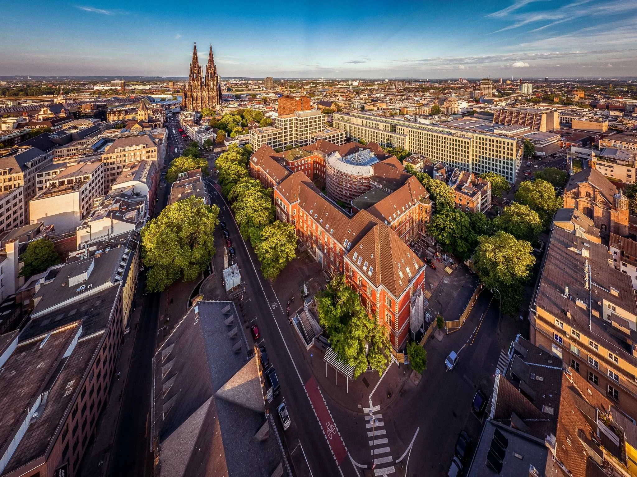 Luftaufnahme des Appellhofplatzes in Köln mit dem historischen Verwaltungsgebäude, dichtem Baumbestand und umliegender Innenstadtbebauung.