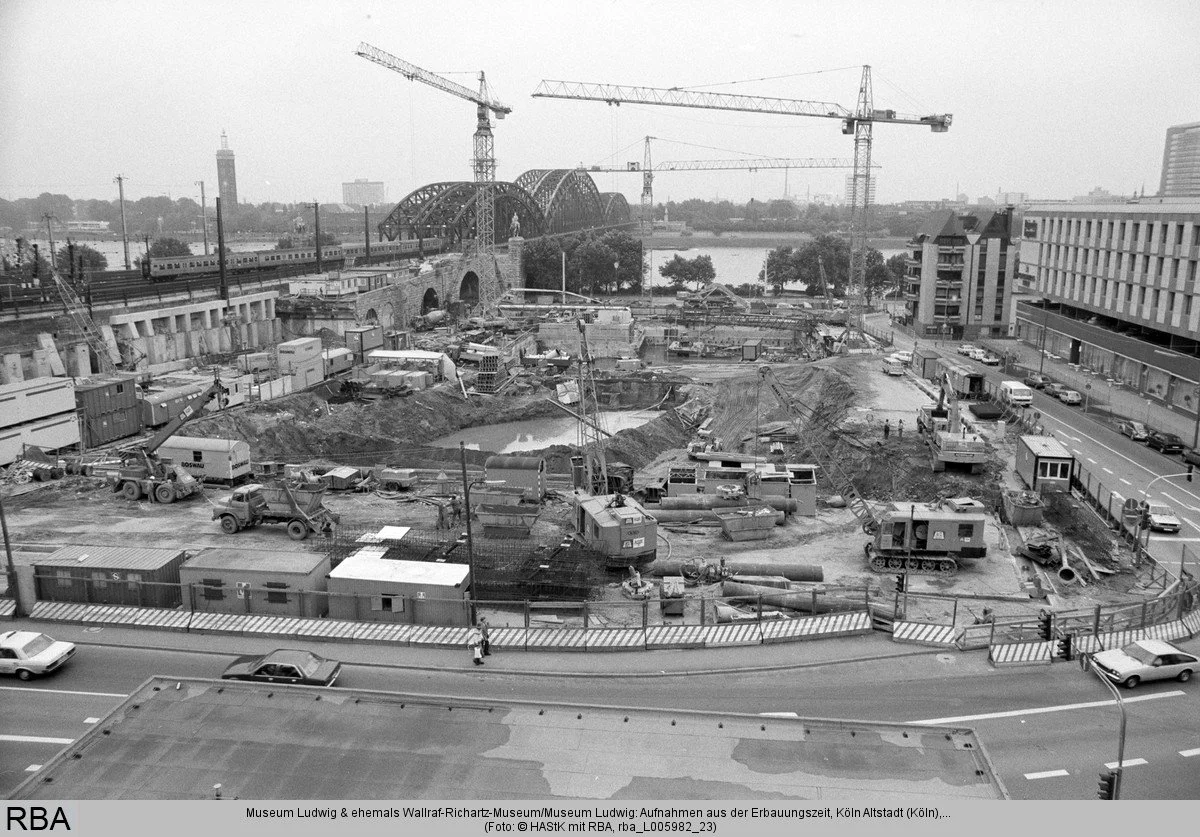 Historische Schwarz-Weiß-Aufnahme der Baustelle von Museum Ludwig und Philharmonie in Köln mit Kränen, Baugruben und der Hohenzollernbrücke im Hintergrund.