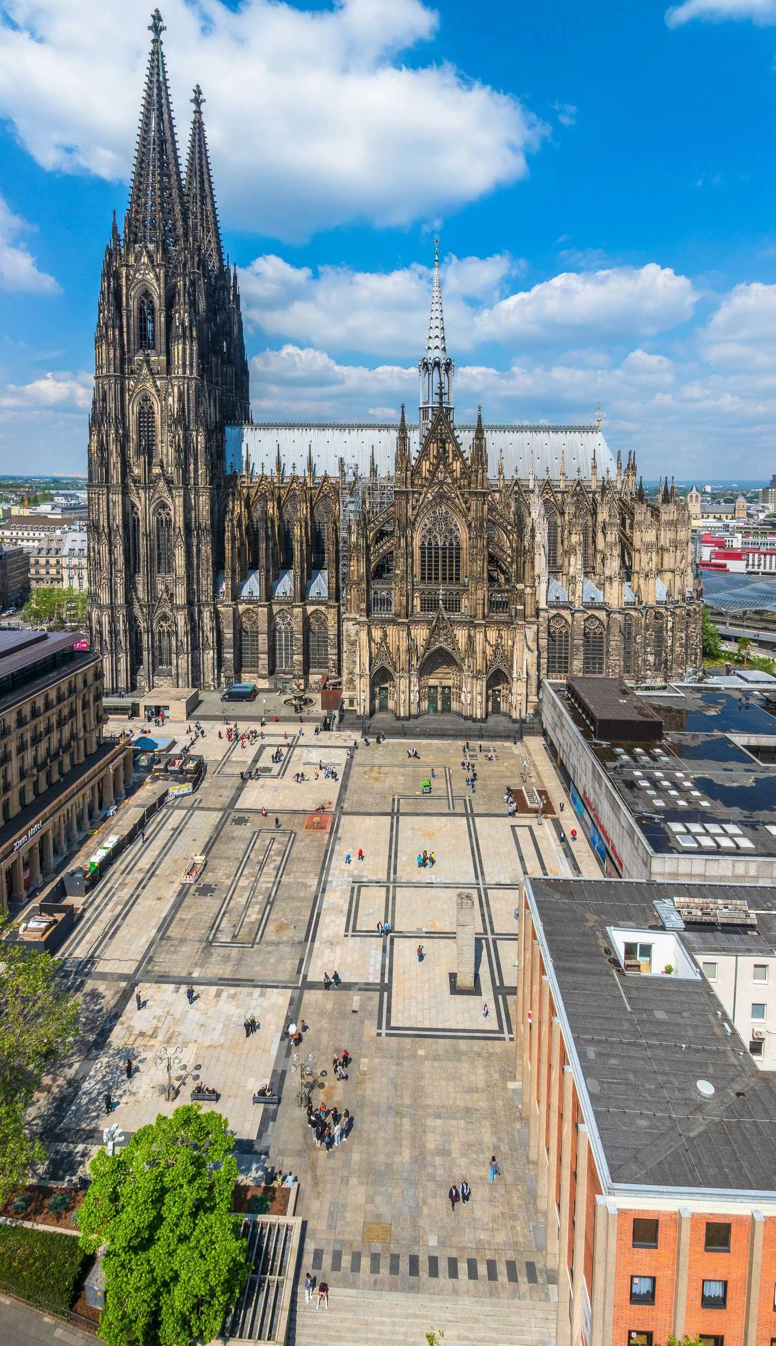 Luftaufnahme des Roncalliplatzes vor dem Kölner Dom bei klarem Himmel. Die beiden Domtürme prägen das Bild, während sich der weitläufige Platz zwischen Dom, Museum Ludwig und den umliegenden Gebäuden erstreckt.