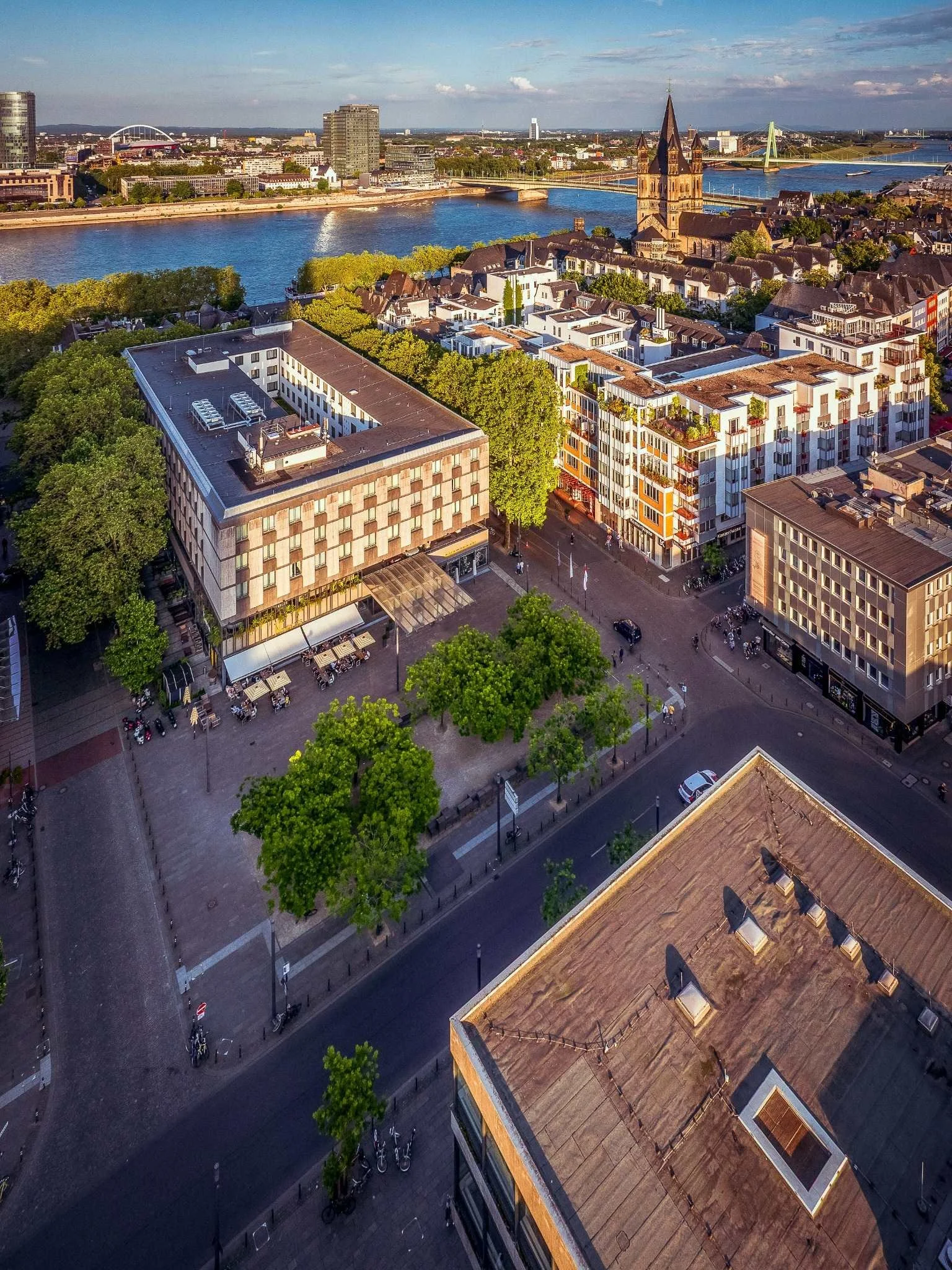Luftaufnahme des Kurt-Hackenberg-Platzes im warmen Abendlicht. Zwischen Museum Ludwig, Philharmonie und umliegenden Gebäuden erstreckt sich der von Bäumen gesäumte Platz; seitlich ist das Museum Ludwig zu sehen.