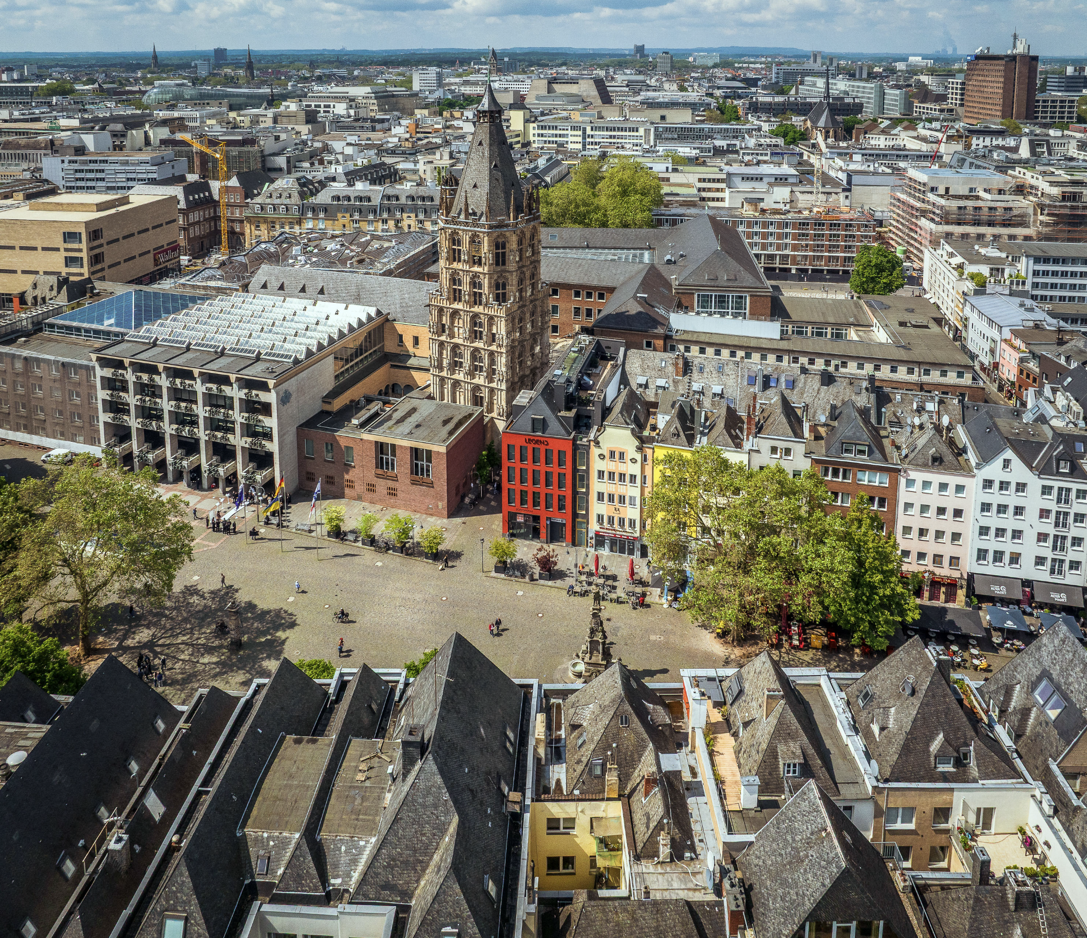 Luftaufnahme des Alter Markts mit dem historischen Rathausturm, bunten Altstadthäusern und dem Jan-von-Werth-Brunnen. Rund um den weitläufigen Platz sind Menschen, Außengastronomie und die dichte Bebauung der Kölner Innenstadt zu sehen.