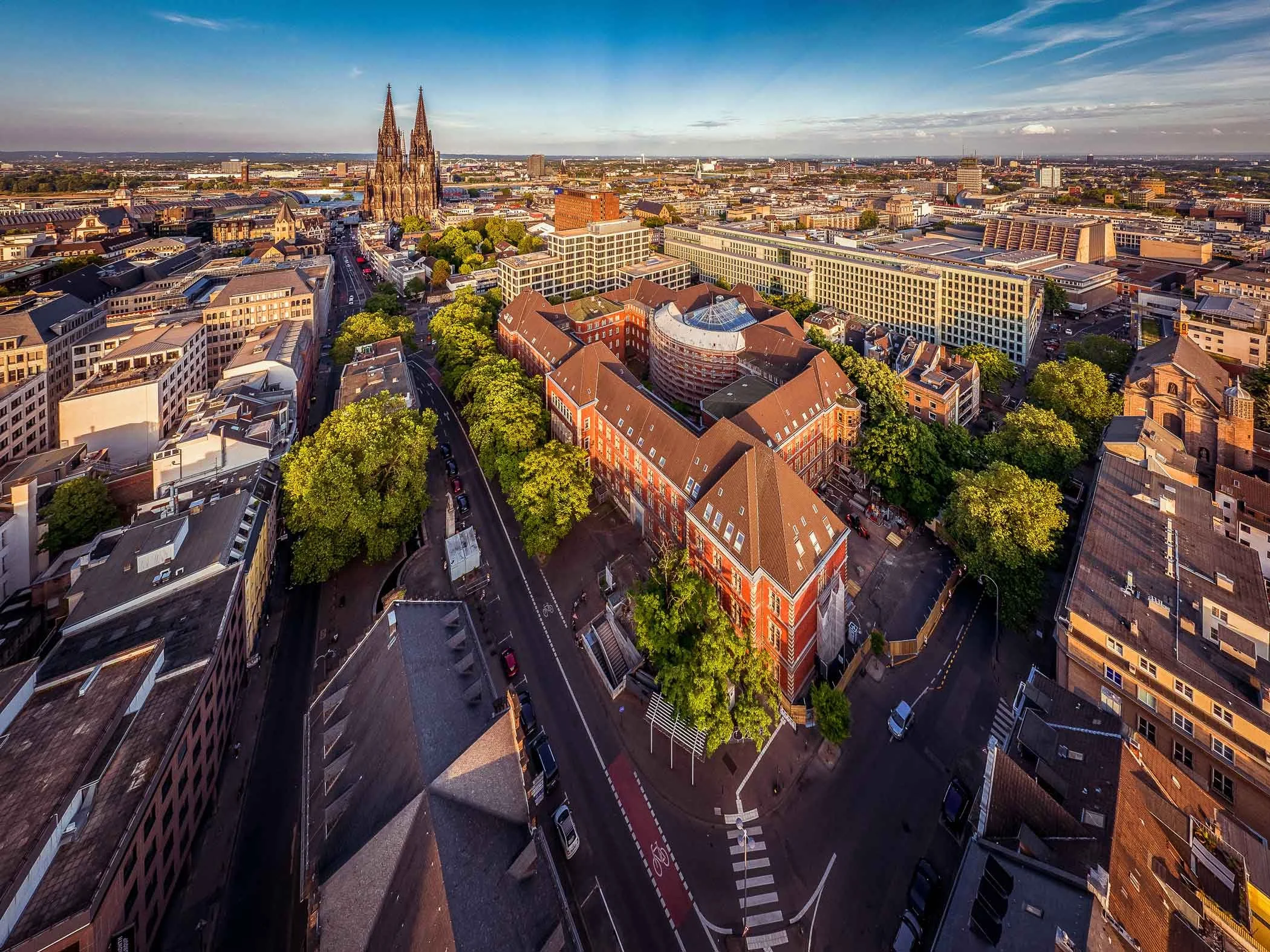 Luftaufnahme des Appellhofplatzes in Köln mit dem historischen Verwaltungsgebäude, dichtem Baumbestand und umliegender Innenstadtbebauung.