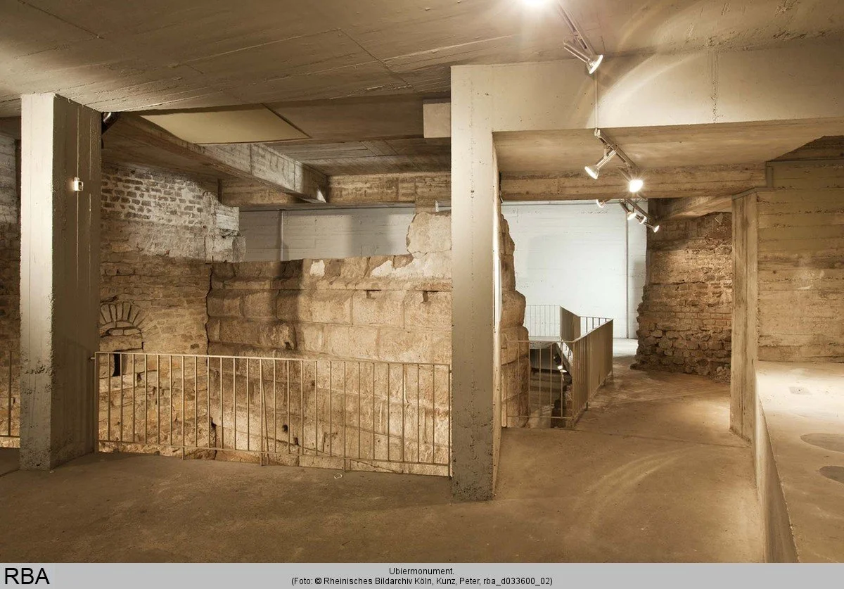 Blick in den unterirdischen Ausstellungsbereich des Ubiermonuments. Zwischen modernen Betonstützen sind die massiven römischen Steinmauern des antiken Bauwerks erhalten und über einen Steg zugänglich.