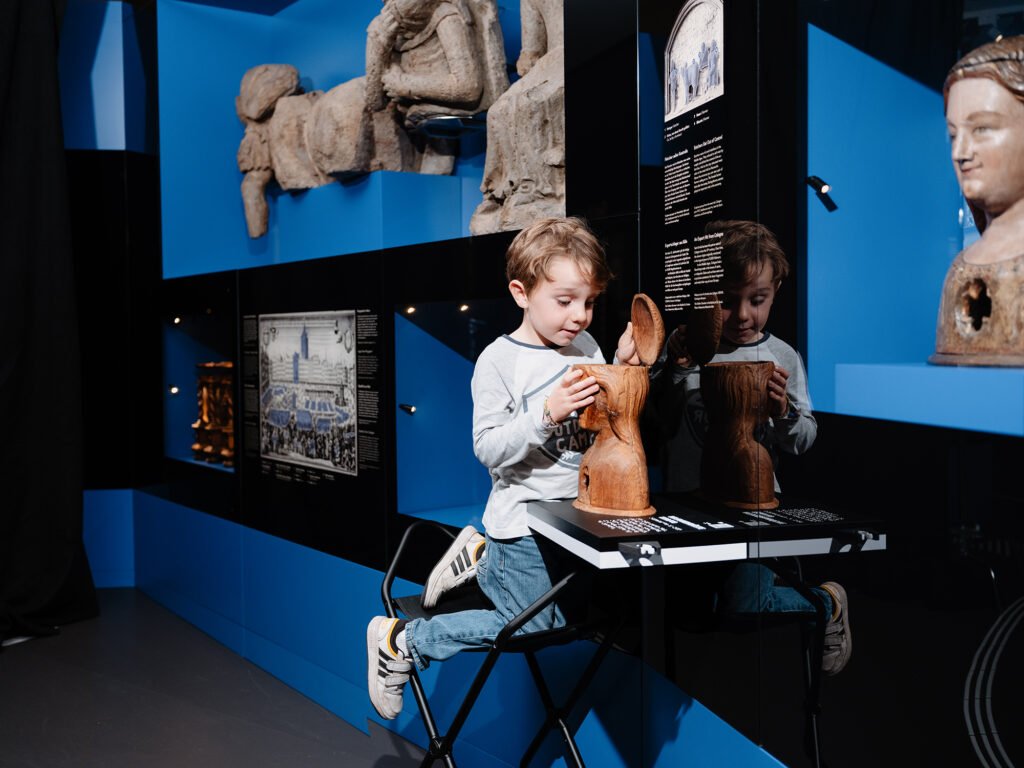Ein Kind erkundet in der interaktiven Dauerausstellung des Kölnischen Stadtmuseums ein hölzernes Ausstellungsobjekt. Im Hintergrund sind historische Skulpturen und Informationstafeln zu sehen.