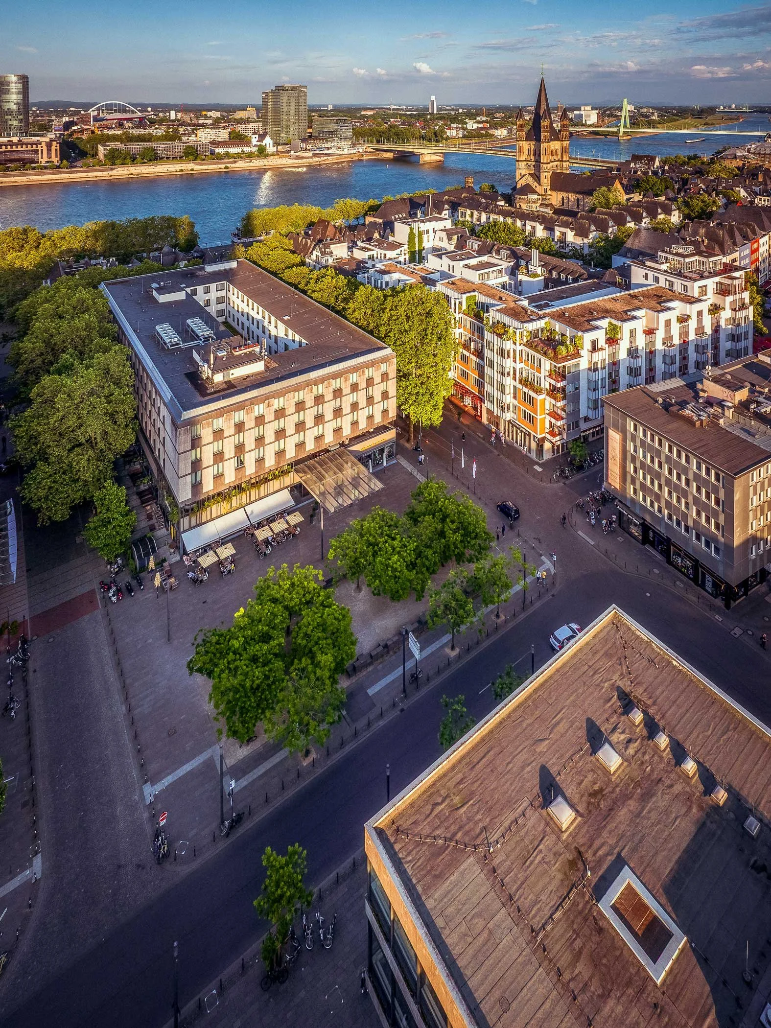 Luftaufnahme des Kurt-Hackenberg-Platzes im warmen Abendlicht. Zwischen Museum Ludwig, Philharmonie und umliegenden Gebäuden erstreckt sich der von Bäumen gesäumte Platz; seitlich ist das Museum Ludwig zu sehen.