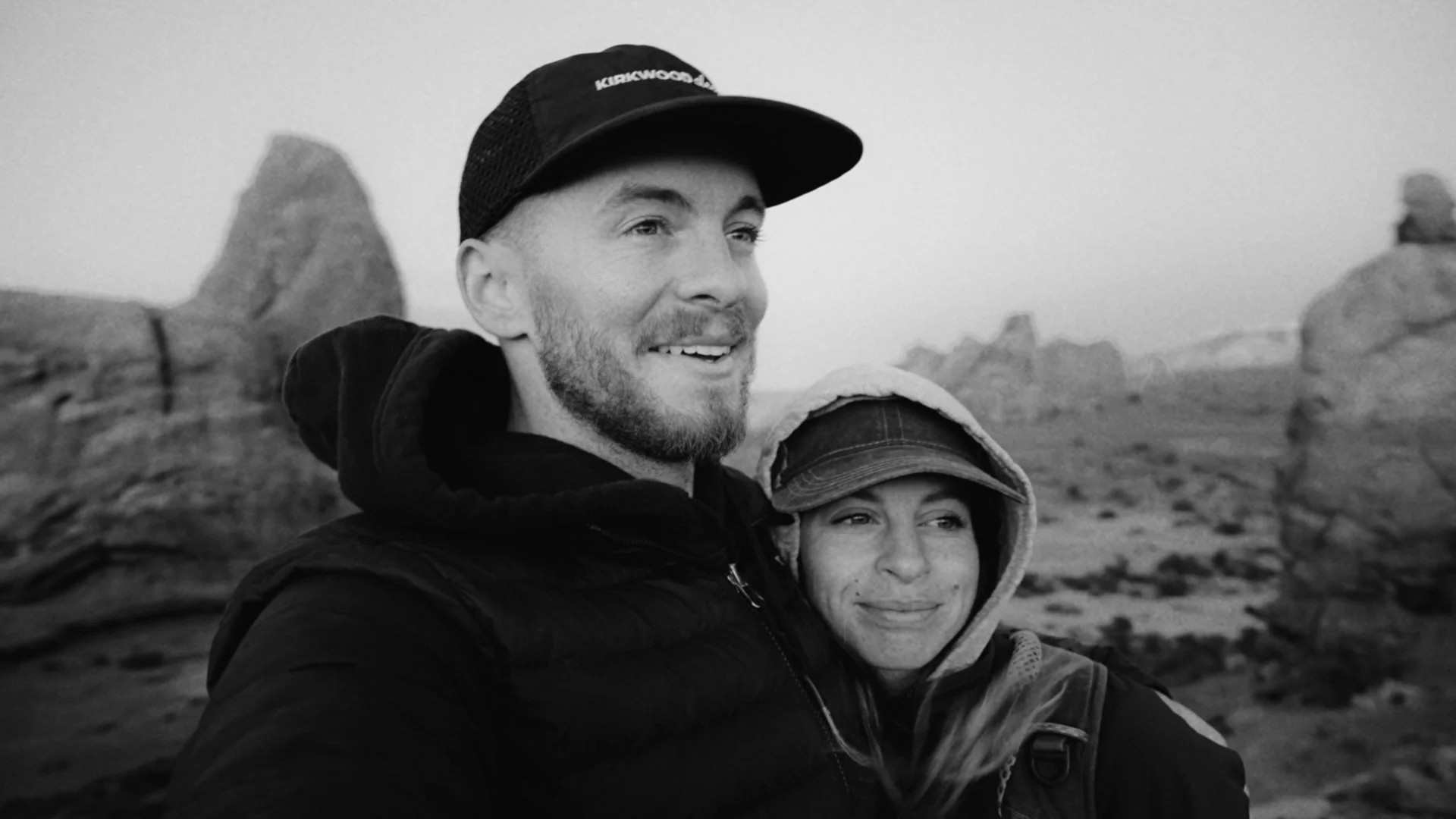 Black and white photo of a couple taking a selfie outdoors with rock formations in the background. The man is smiling, wearing a cap and a jacket, and the woman is smiling as well, wearing a hooded jacket and a cap.