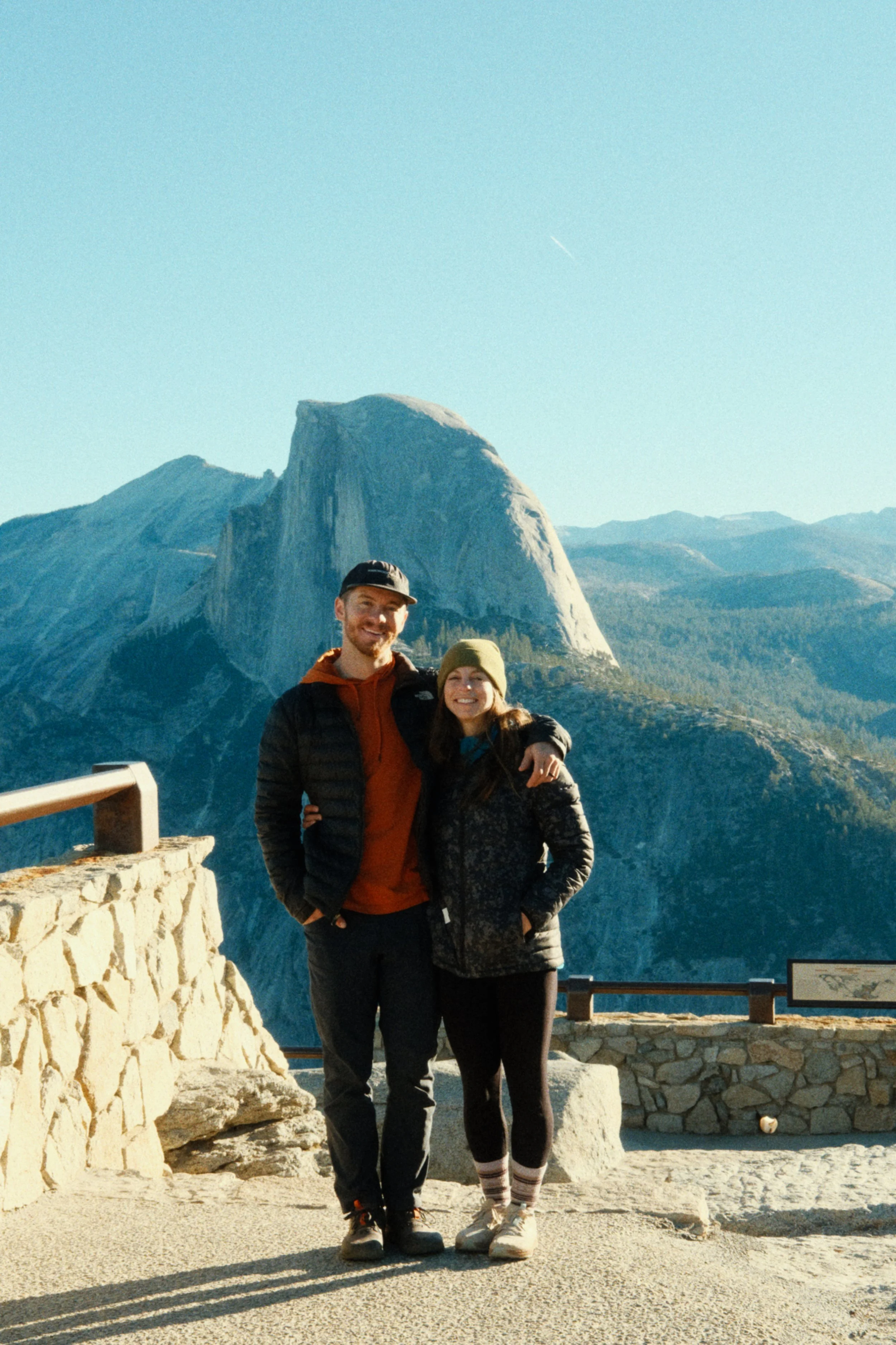 A couple stands close together, smiling, with Yosemite National Park's Half Dome and surrounding mountains in the background.