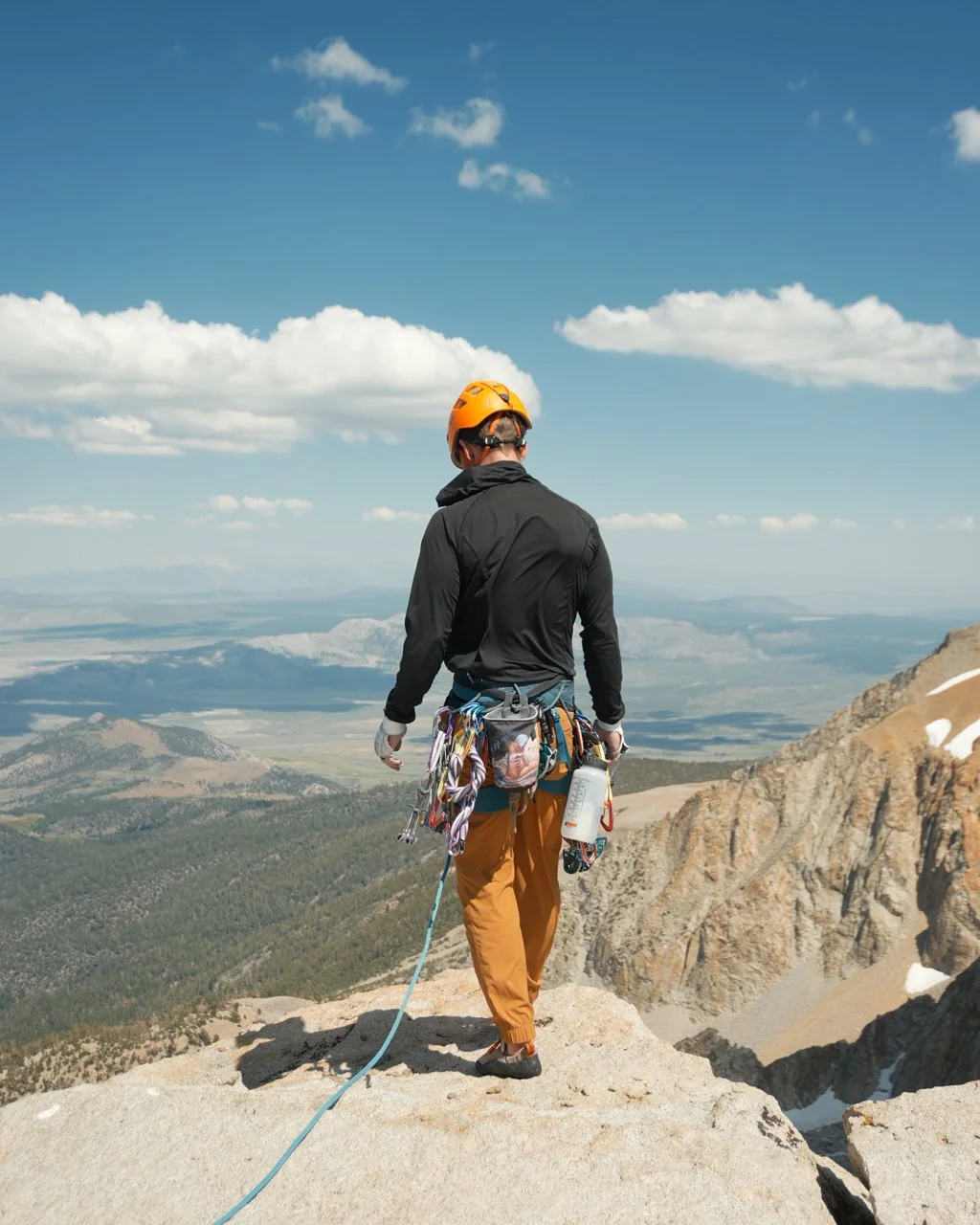A person wearing a yellow helmet and black jacket standing on a rocky mountain summit with climbing gear, overlooking a vast landscape of mountains and valleys under a partly cloudy sky.