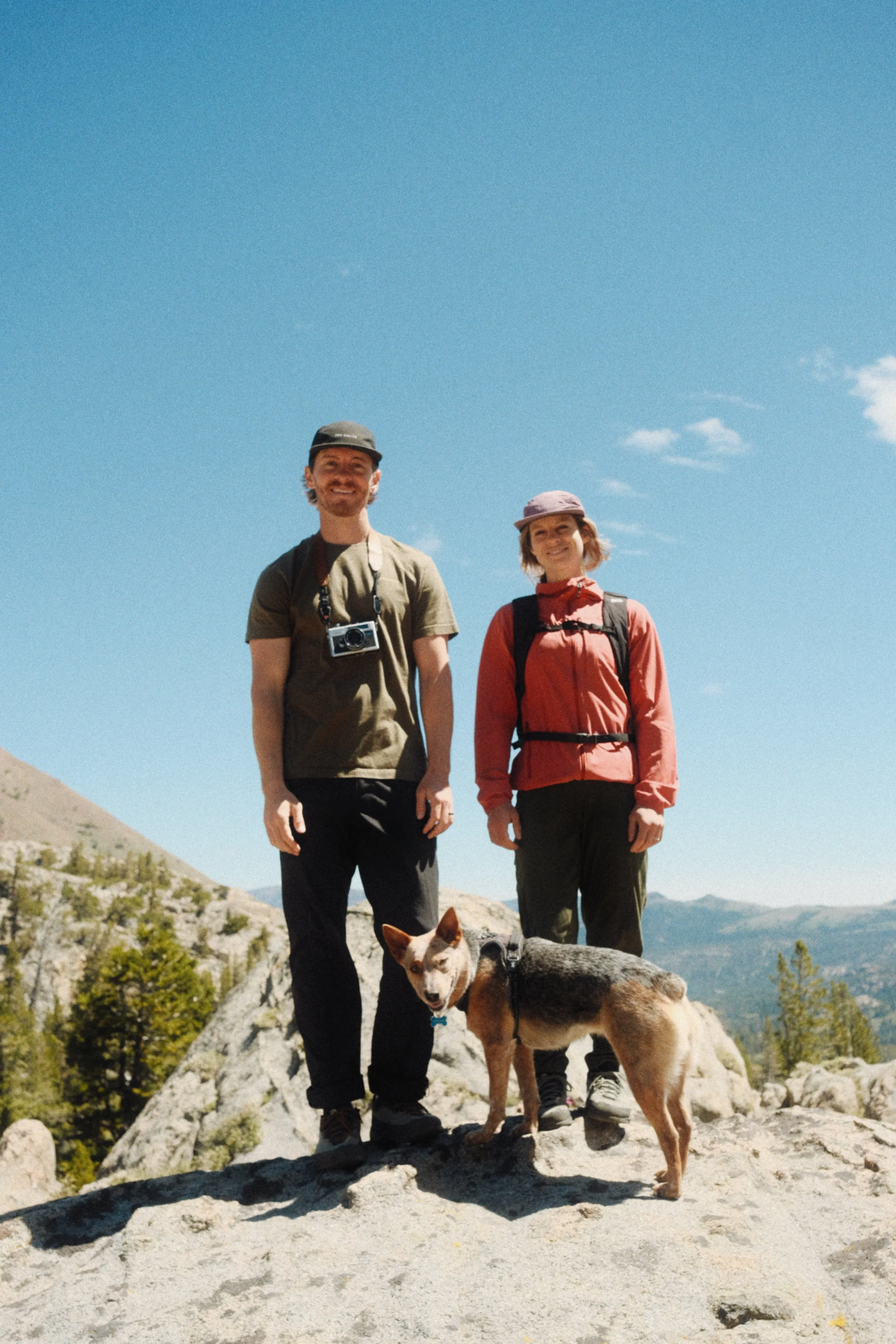 Two hikers, a man and a woman, standing on rocky terrain with a scenic mountain landscape and blue sky in the background, accompanied by a dog.