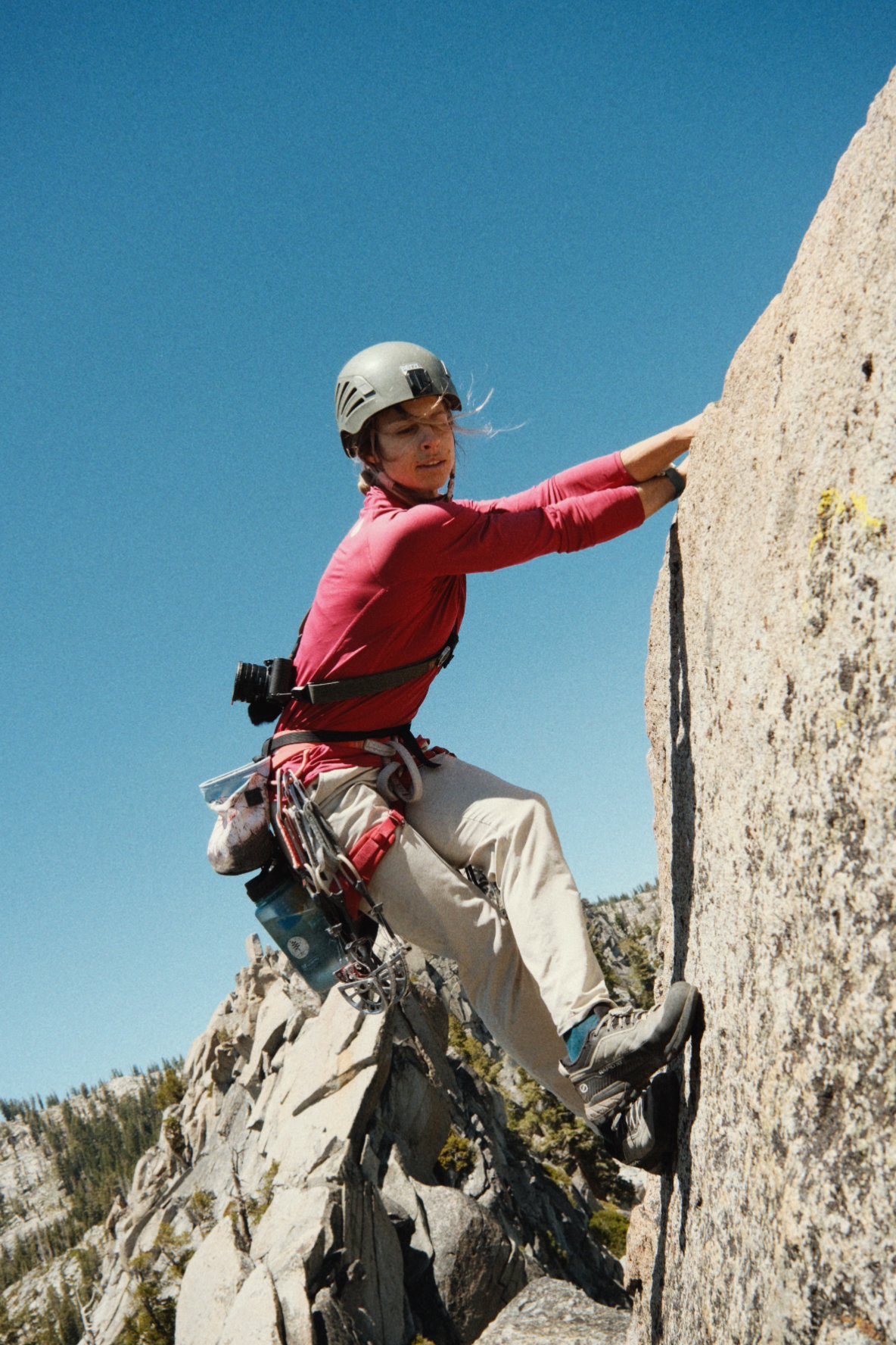 A woman rock climbing outdoors on a steep rock face, wearing a helmet, climbing harness, and outdoor gear.
