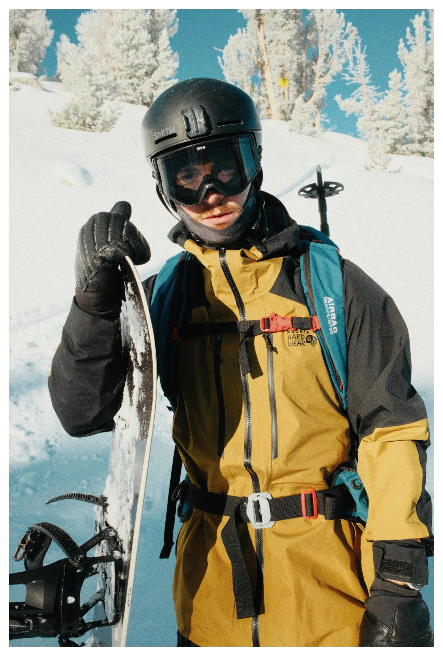 A man dressed in yellow and black winter gear, wearing a helmet and goggles, holding skis against a snowy landscape with snow-covered trees and a clear blue sky.