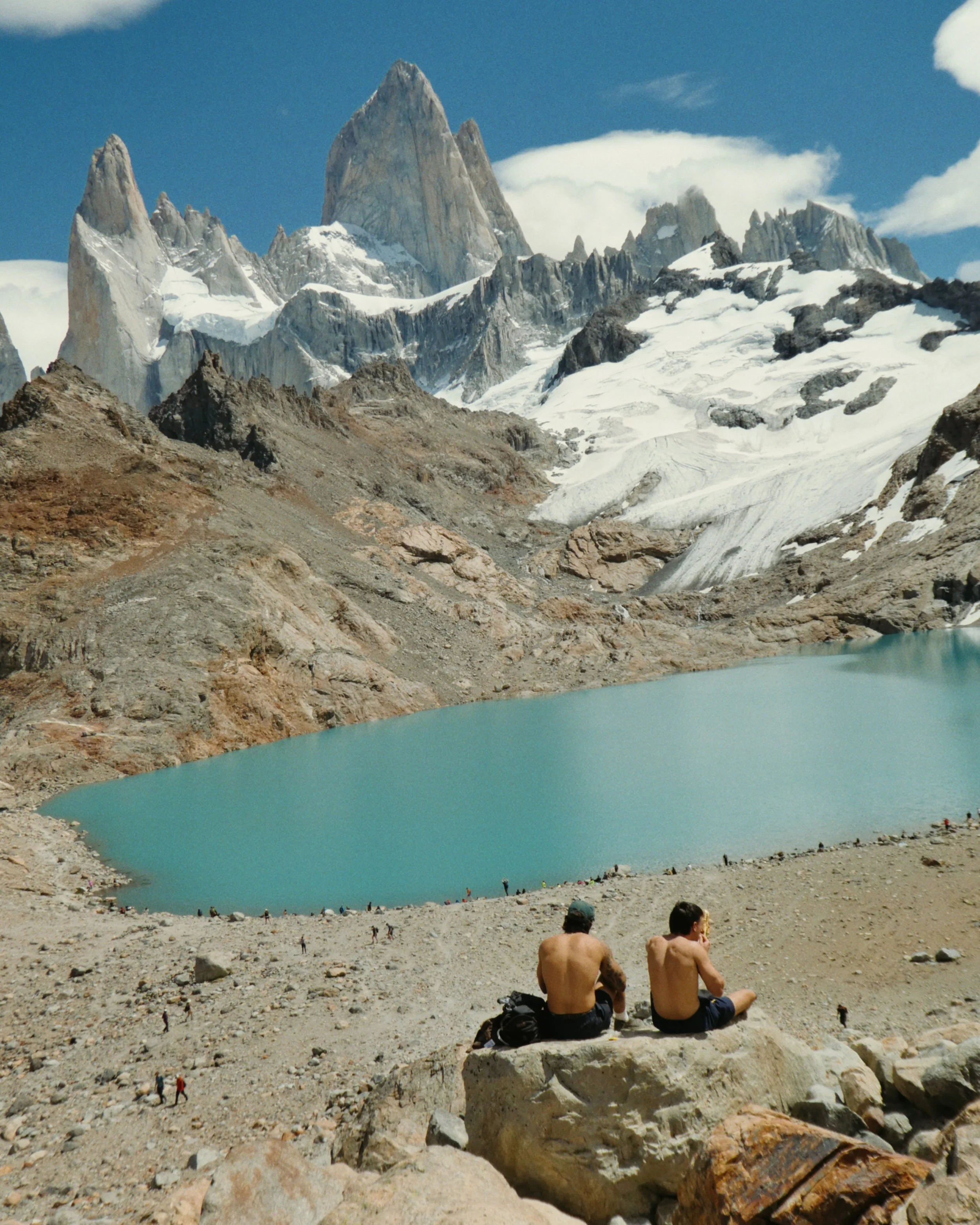 A view of a glacial mountain landscape with rugged peaks, snow, and a turquoise glacial lake. In the foreground, two shirtless men sit on rocks, facing away from the camera, with several small people walking around the rocky terrain.