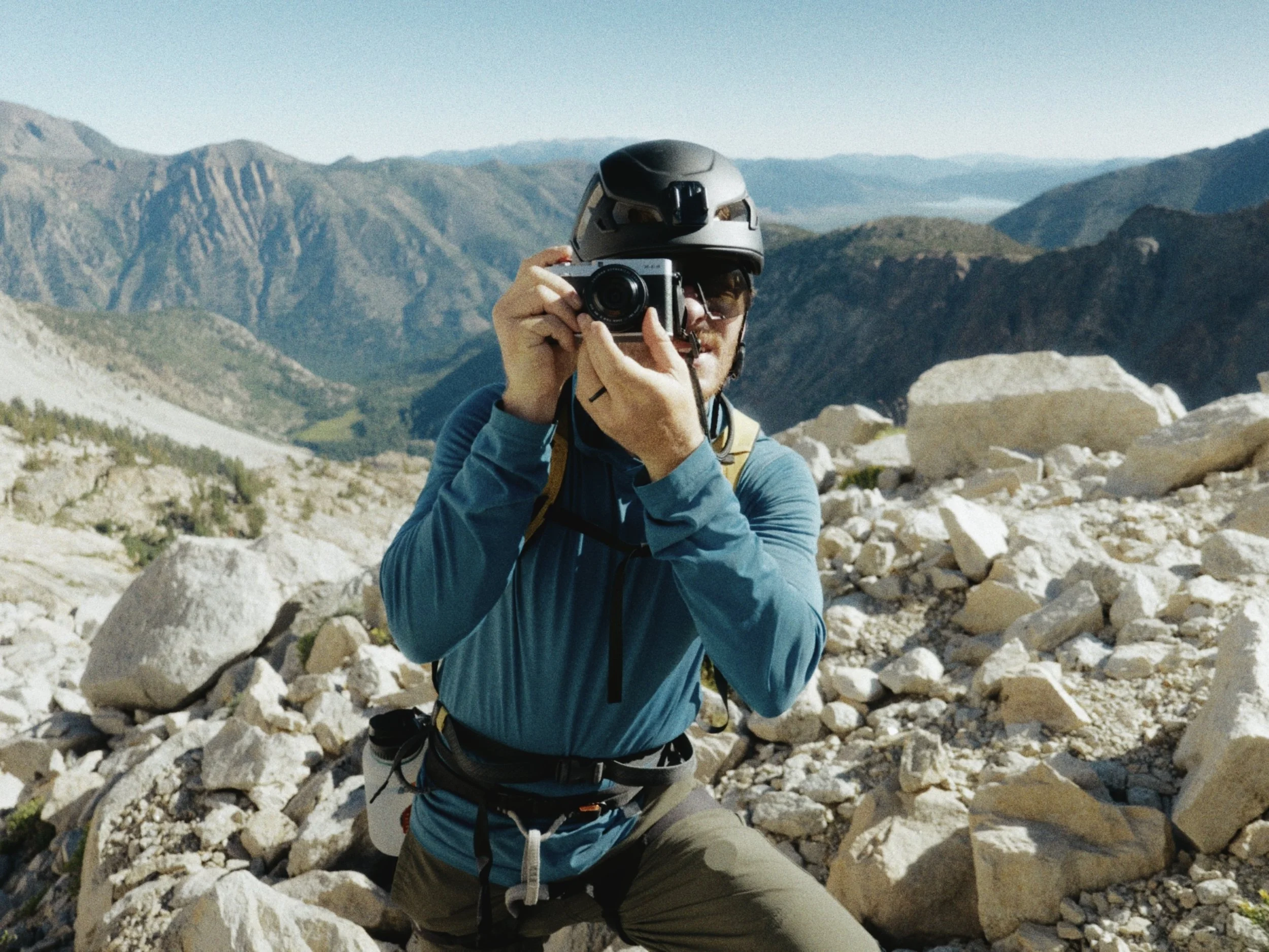 A person taking a photograph with a camera while hiking in a rocky, mountainous landscape wearing a helmet, glasses, and outdoor clothing.