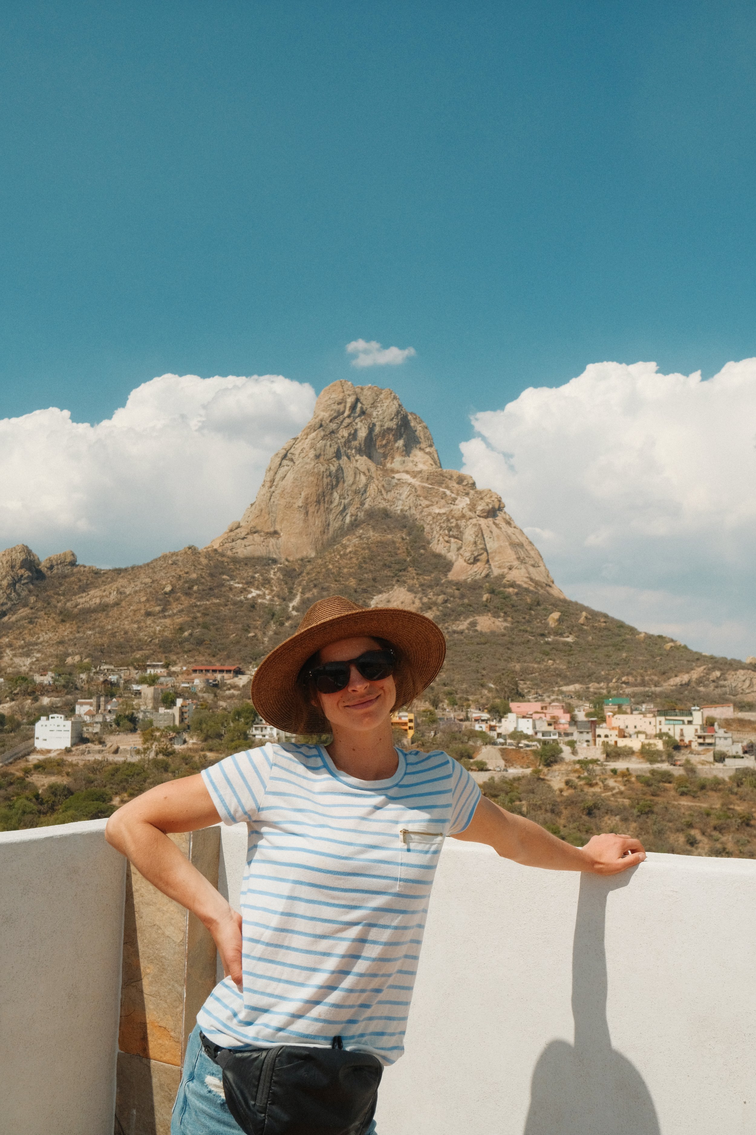 A smiling woman with sunglasses and a wide-brimmed hat standing outdoors with a mountain and town in the background.