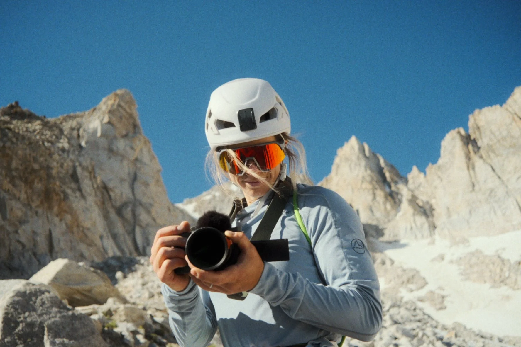 A person in outdoor mountaineering gear, including a white helmet and orange reflective goggles, holding a camera in a snowy mountainous landscape with rocky peaks and a clear blue sky.