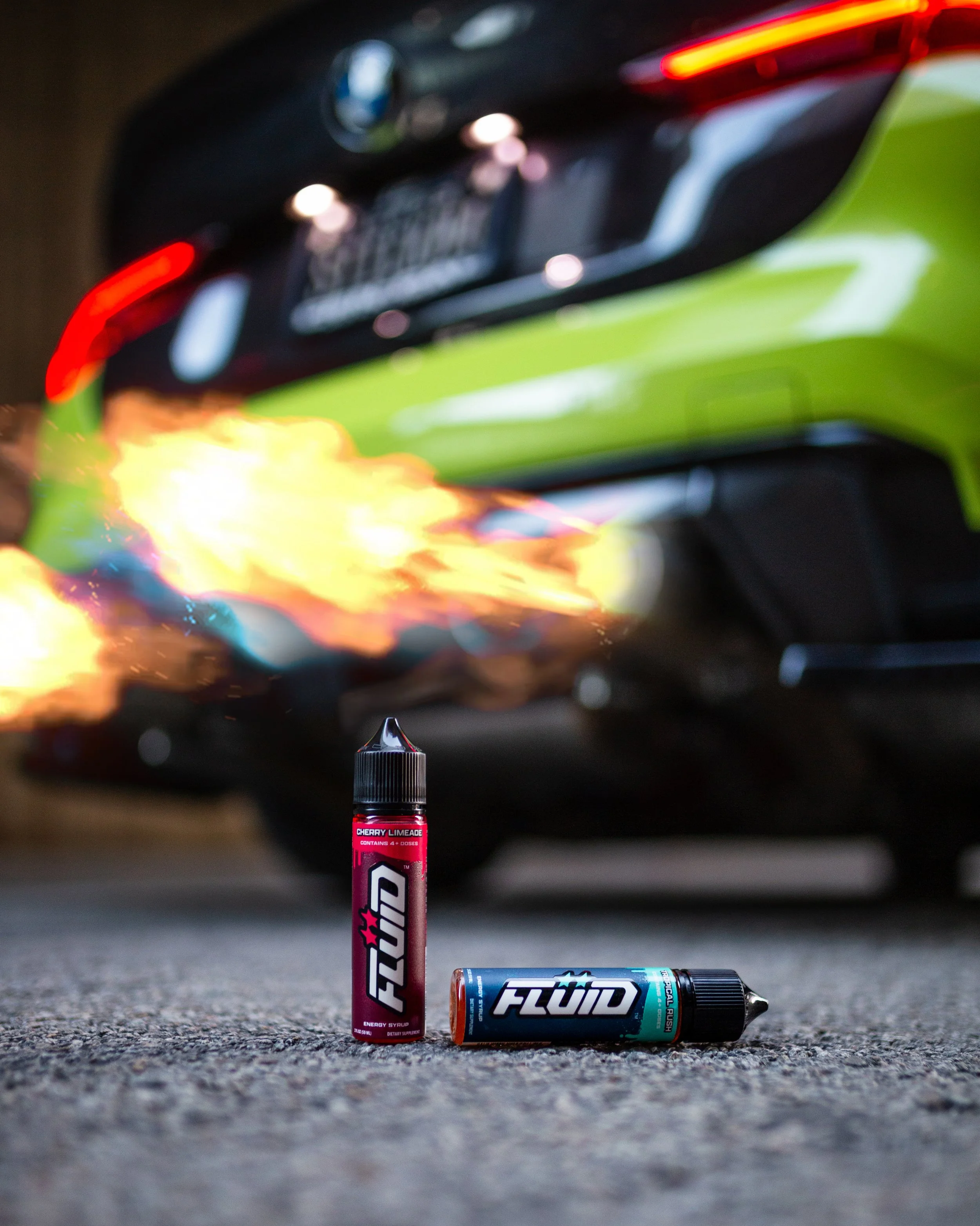 FLUID Energy Syrup bottles on pavement with flames shooting from a lime-green sports car’s exhaust in the background