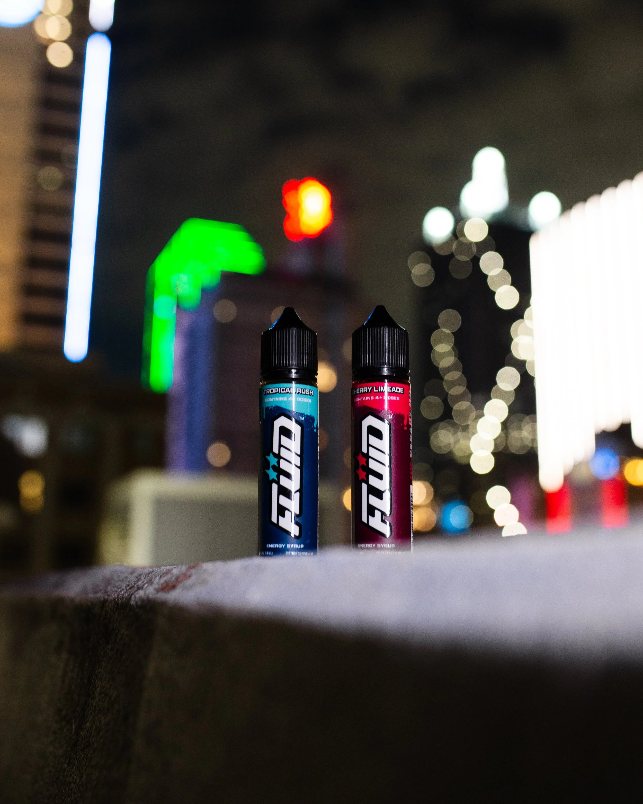 Two FLUID Energy Syrup bottles standing on a concrete ledge with blurred Dallas skyline lights in the background at night