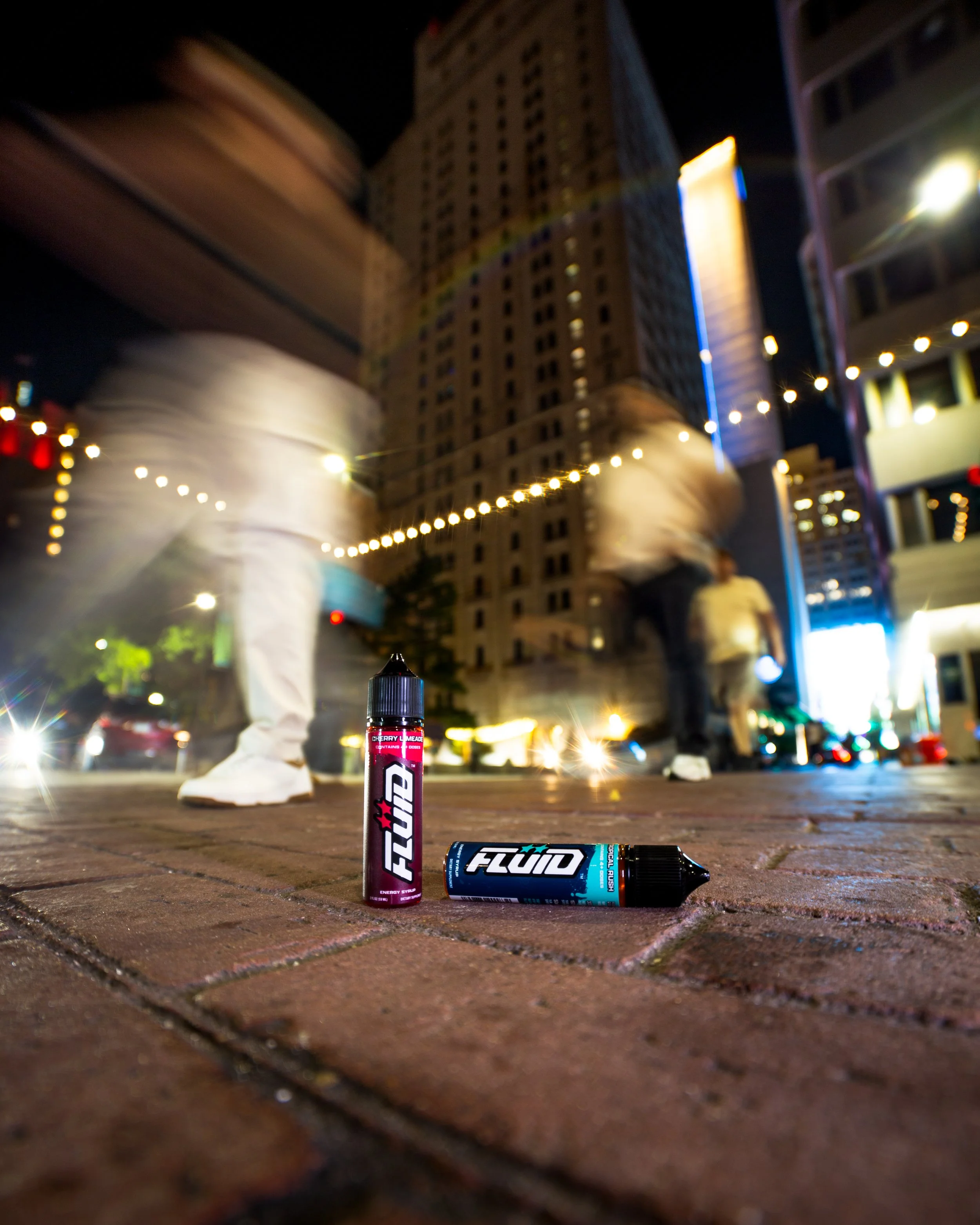 FLUID Energy Syrup bottles on a downtown sidewalk at night with motion-blurred pedestrians and illuminated buildings in the background