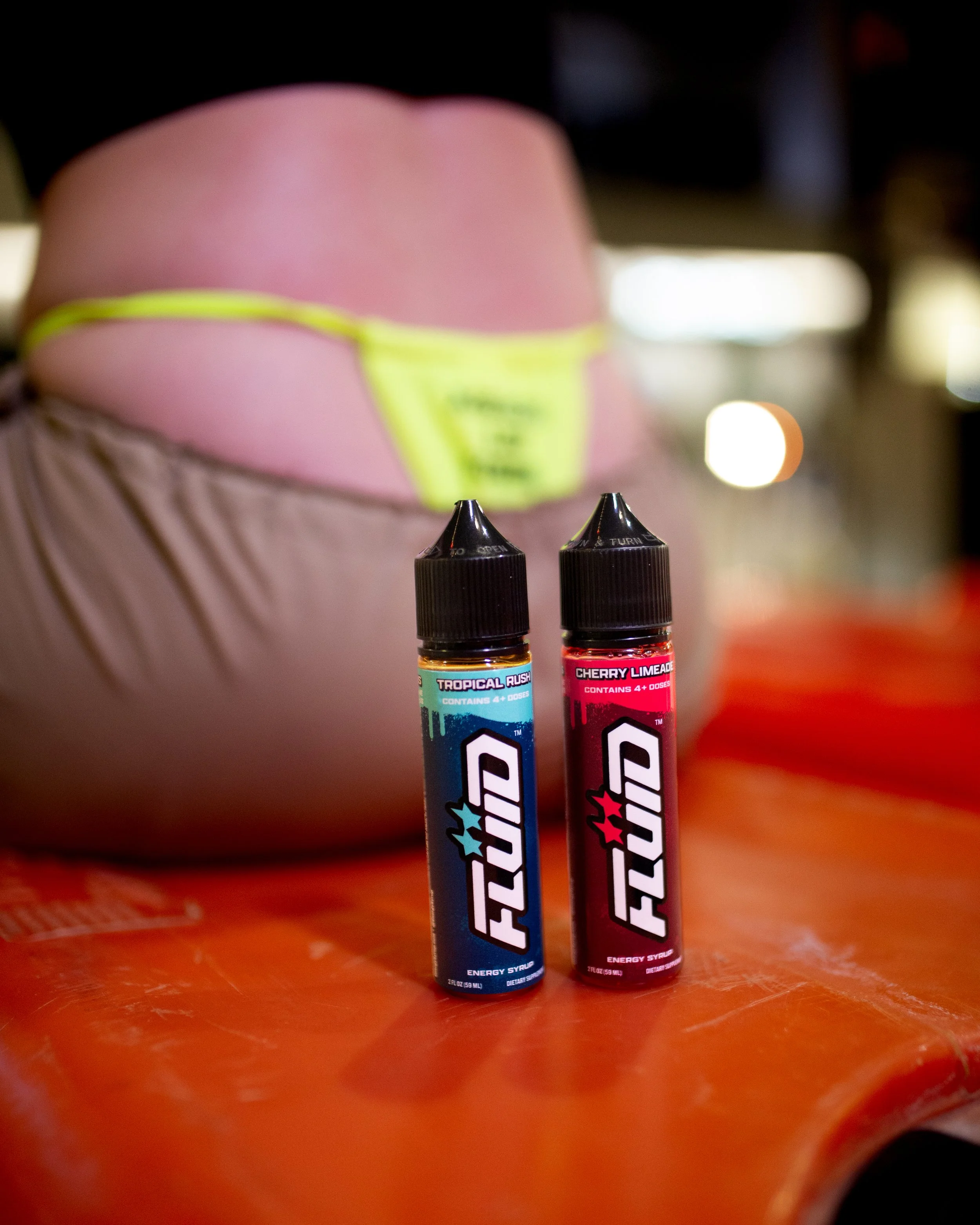 Two FLUID Energy Syrup bottles on an orange surface with a person wearing a neon-green thong in the background