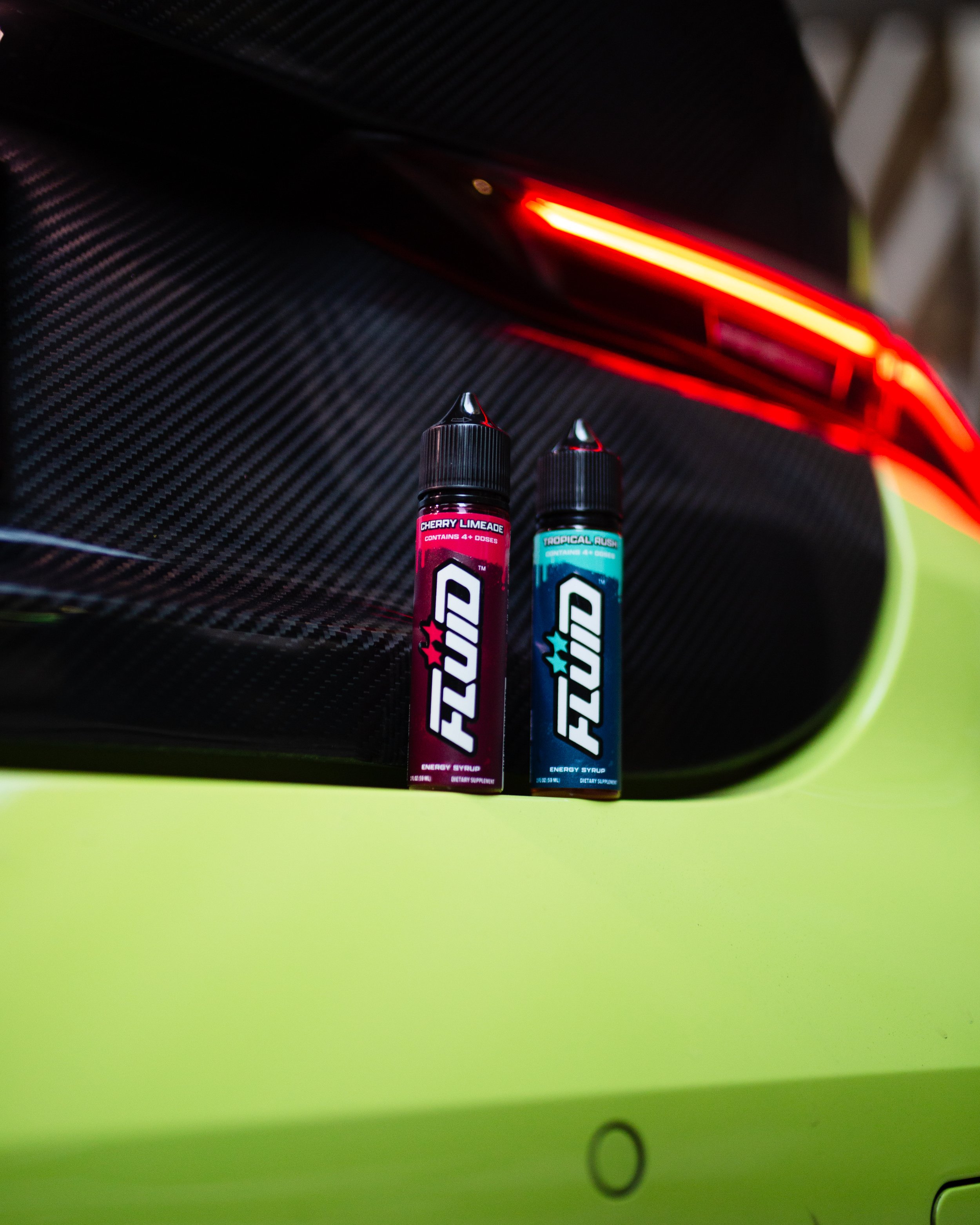 Two FLUID Energy Syrup bottles against a lime-green sports car’s carbon-fiber rear panel beneath a red taillight