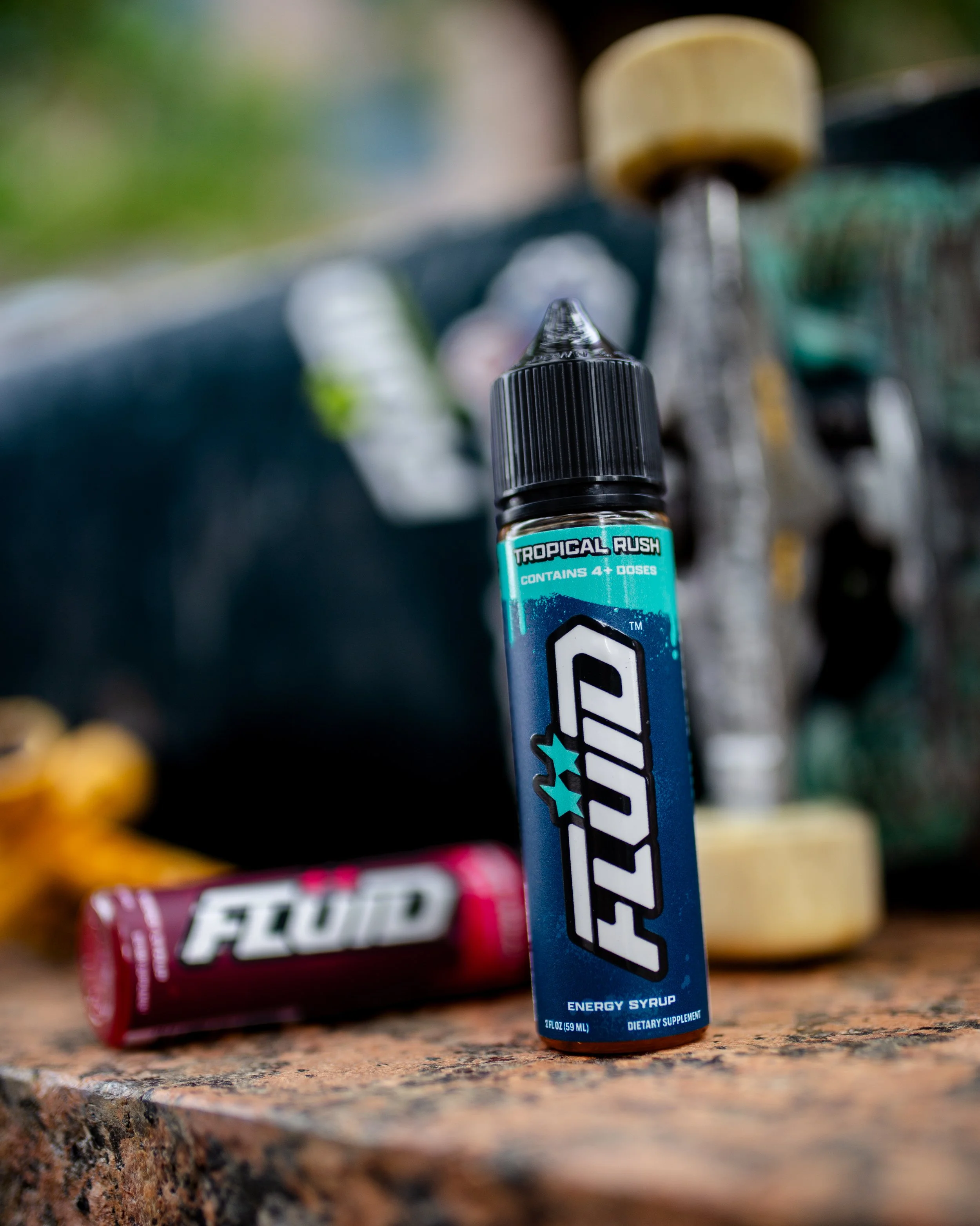 Blue-labeled FLUID Energy Syrup bottle standing beside a red-labeled bottle with a worn skateboard in the background