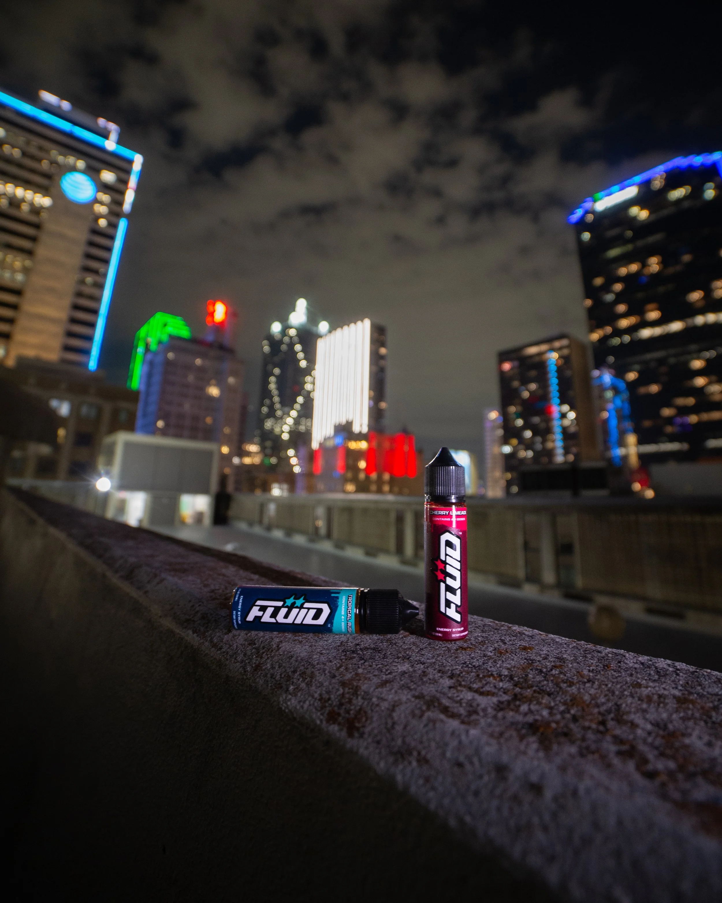 FLUID Energy Syrup bottles on a concrete ledge overlooking the illuminated Dallas skyline at night