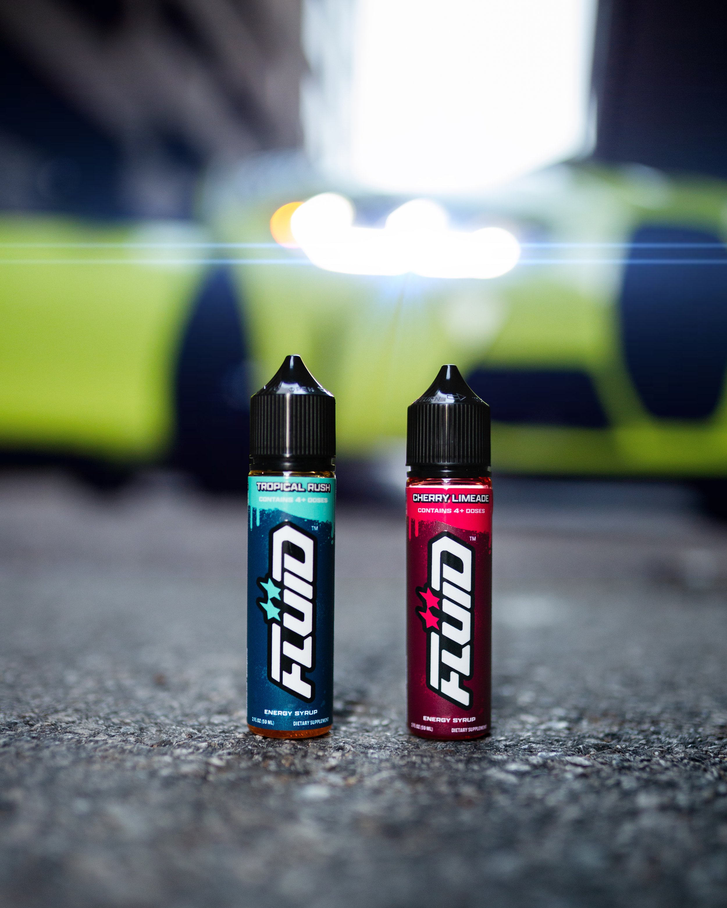 Two FLUID Energy Syrup bottles on pavement with a lime-green sports car in the background