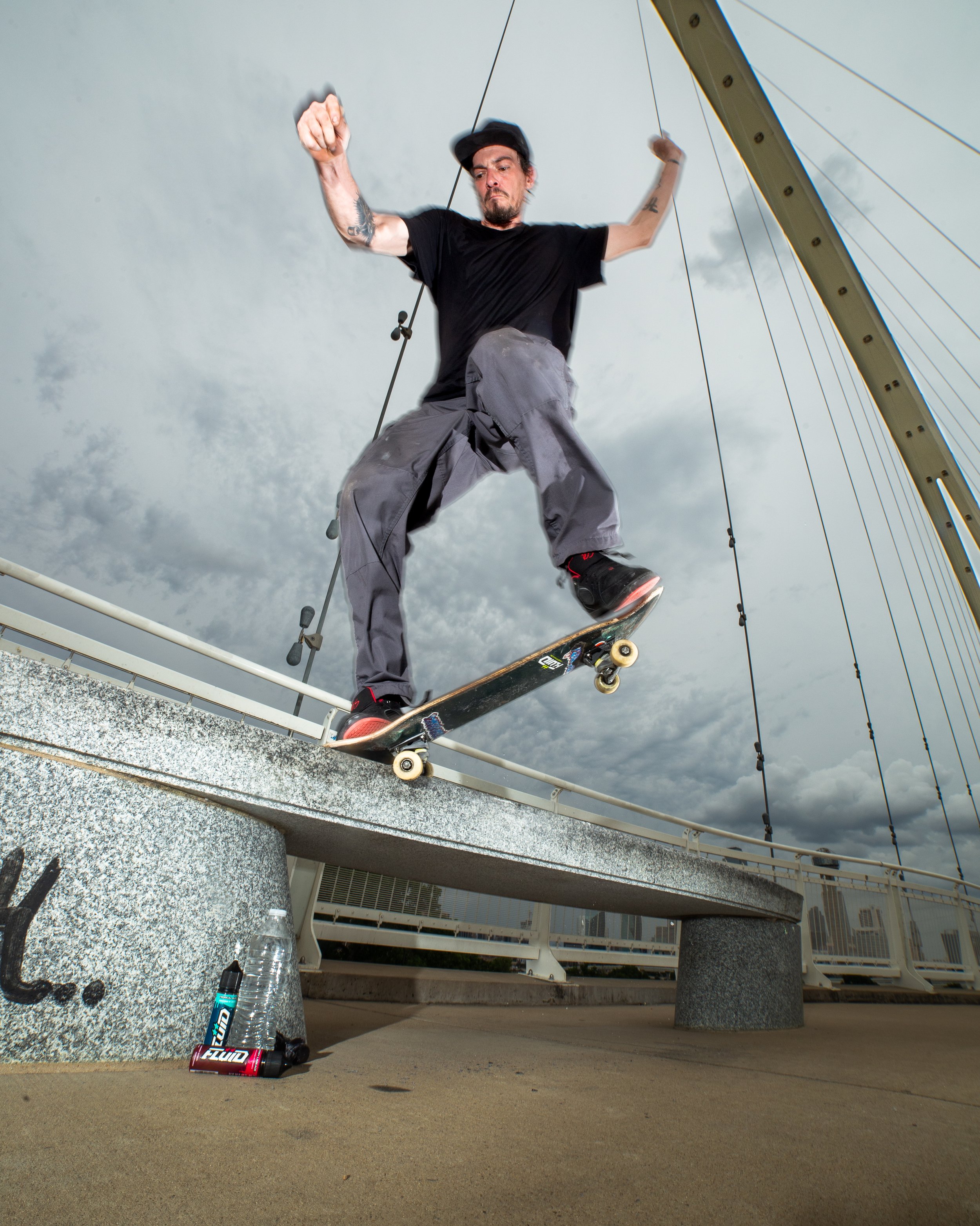 Skateboarder performing a rail trick beneath a bridge with FLUID Energy Syrup bottles on the concrete below