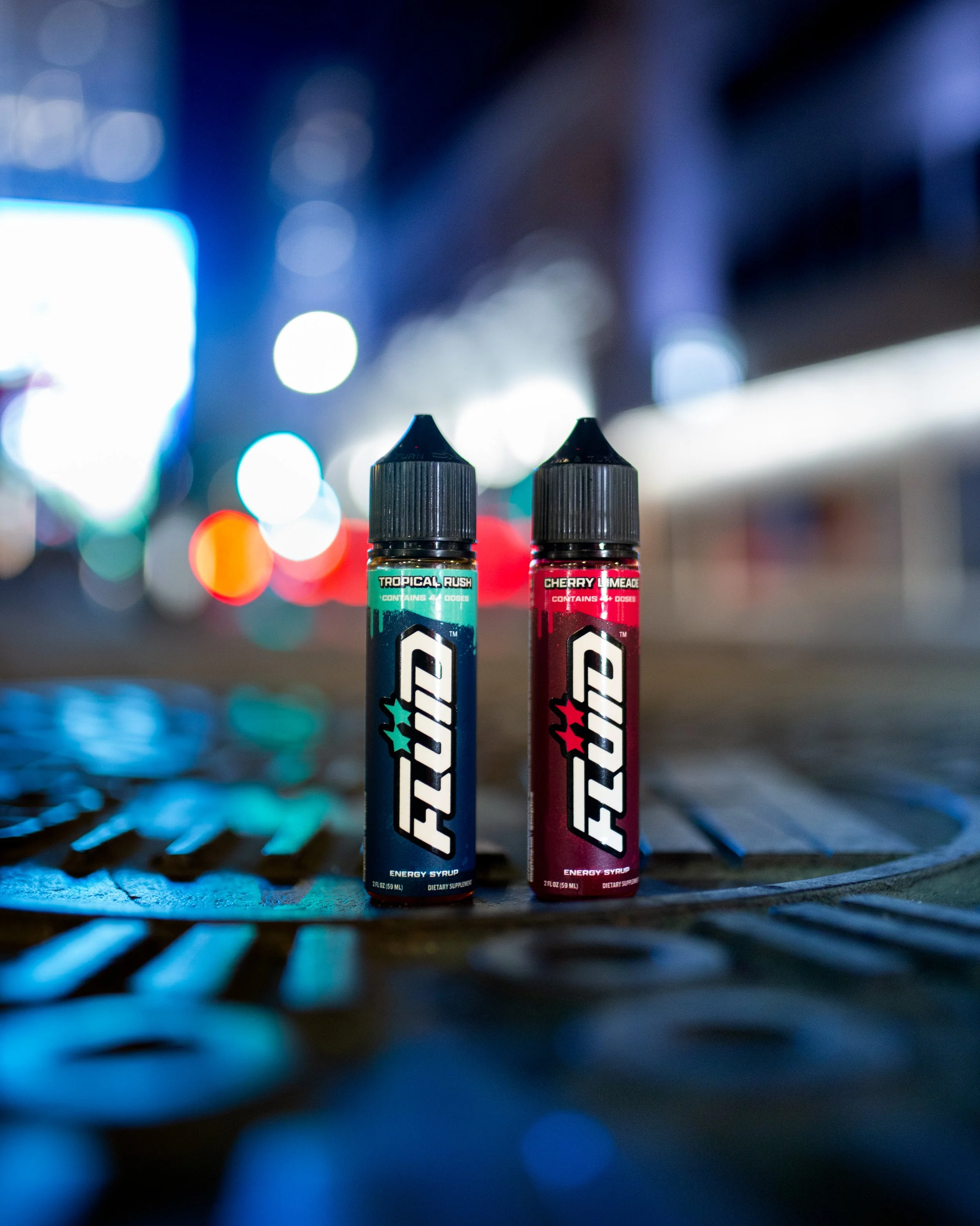 Two FLUID Energy Syrup bottles standing on a patterned metal surface with blurred city lights in the background at night
