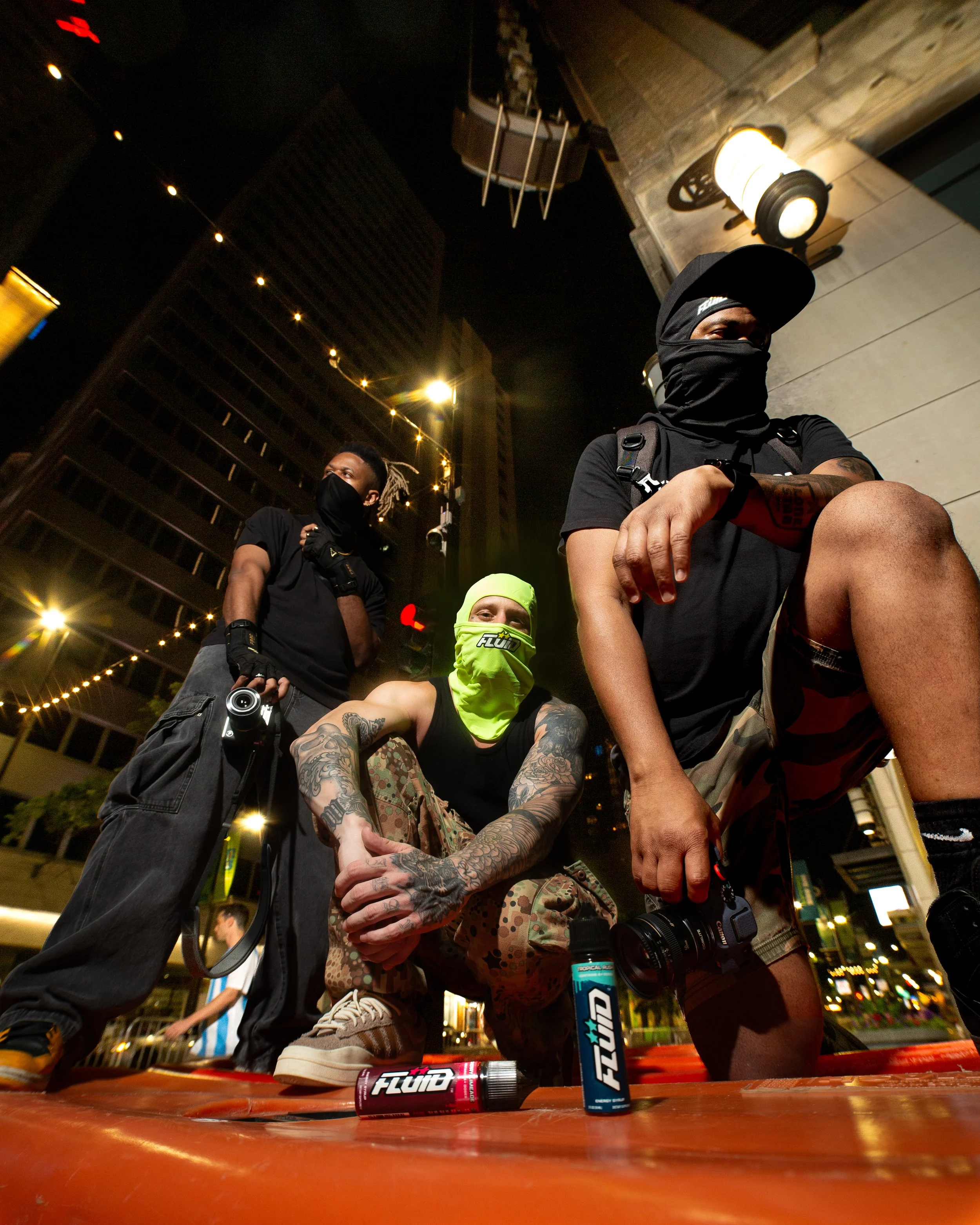 Three people posing beneath city lights at night with FLUID Energy Syrup bottles in the foreground
