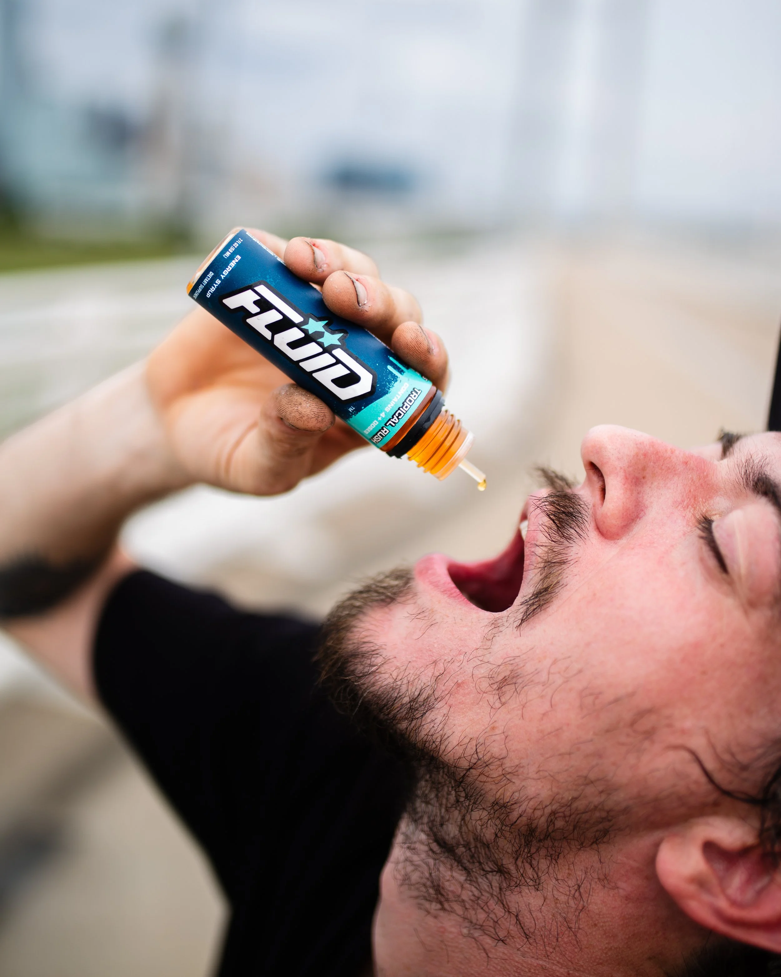 Close-up of a person dispensing FLUID Energy Syrup from a blue-labeled bottle into their mouth outdoors