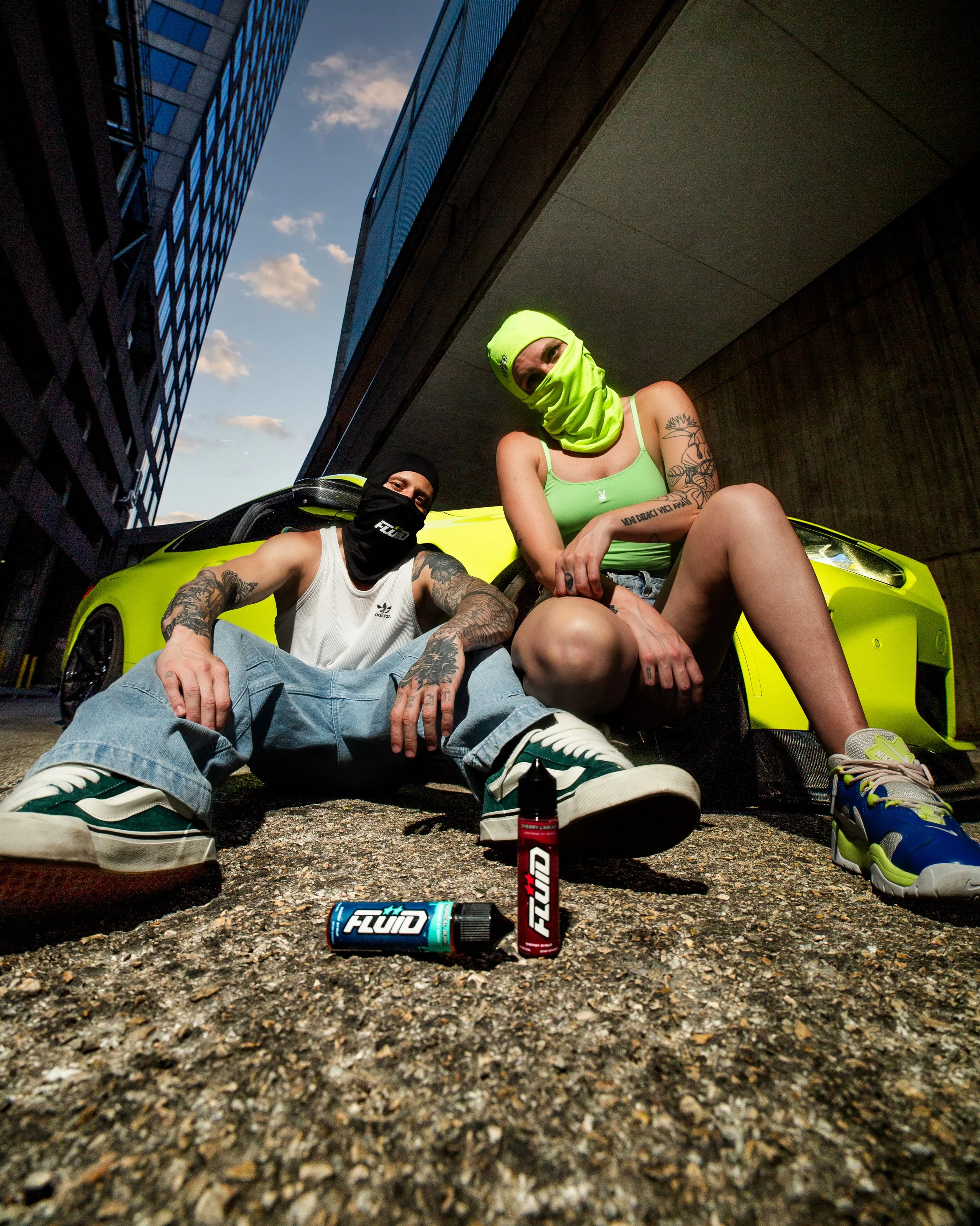 Two people posing beside a lime-green sports car beneath an overpass with FLUID Energy Syrup bottles in the foreground