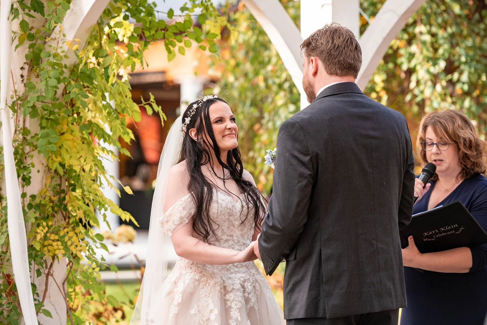 Bride and groom exchanging vows during an outdoor wedding ceremony, with a officiant reading from a book, standing under a white arch decorated with green vines.