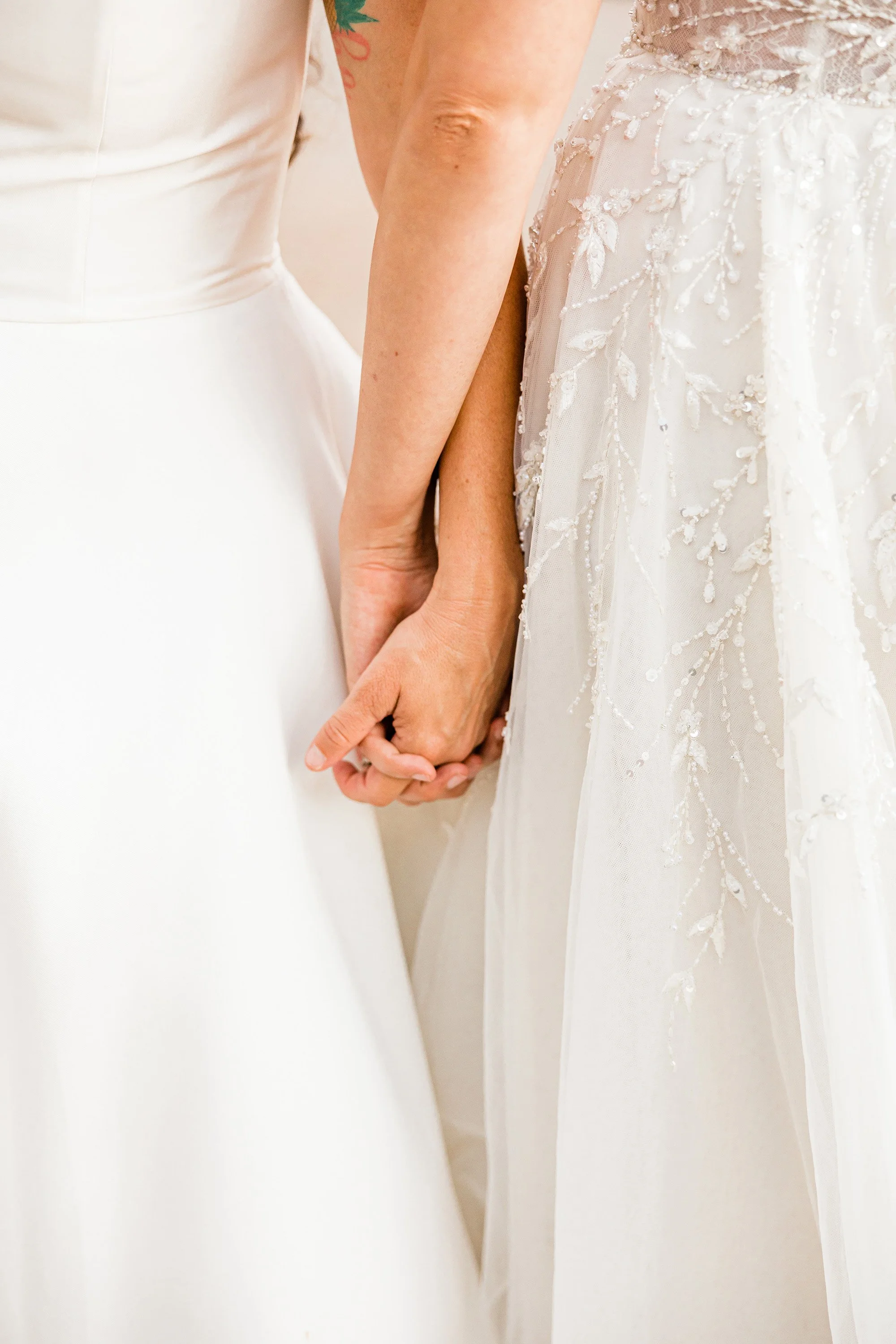 Two women holding hands, wearing wedding dresses.