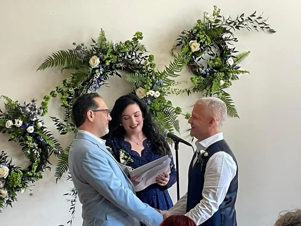 Two men holding hands, presumably at a wedding ceremony, with a woman officiating. Floral arrangements decorate the background.