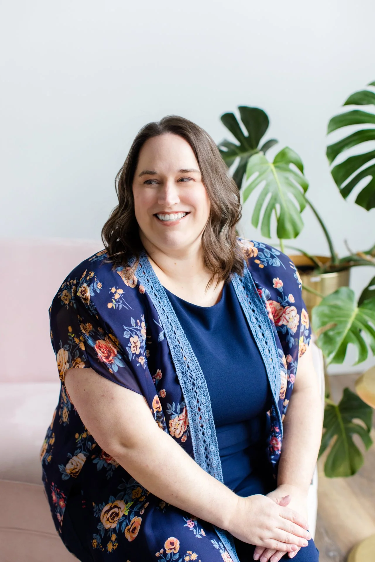 A woman with shoulder-length brown hair, wearing a dark blue dress with floral accents and a matching floral cardigan, sitting on a light pink sofa in front of a large green plant, smiling and looking to her left.
