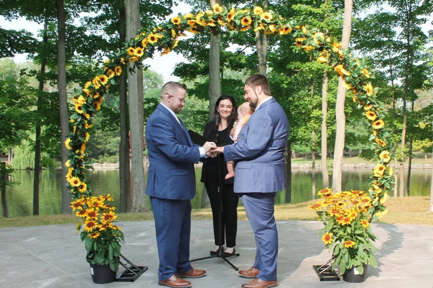 Two grooms holding hands under a sunflower arch in front of an officiant