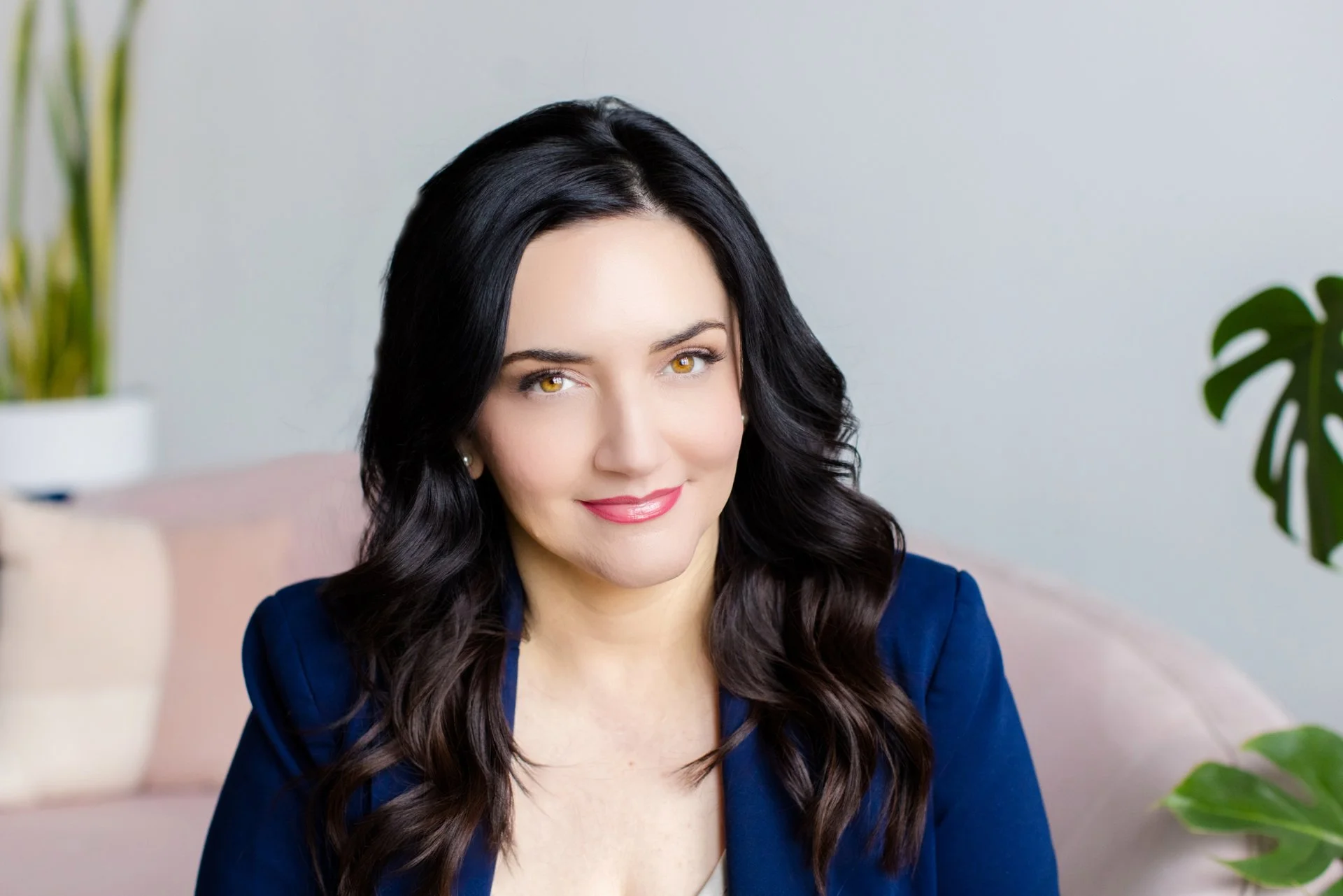 Portrait of a woman with long dark wavy hair, wearing a blue blazer, sitting on a light-colored sofa in a cozy, modern living room with indoor plants and a light background.