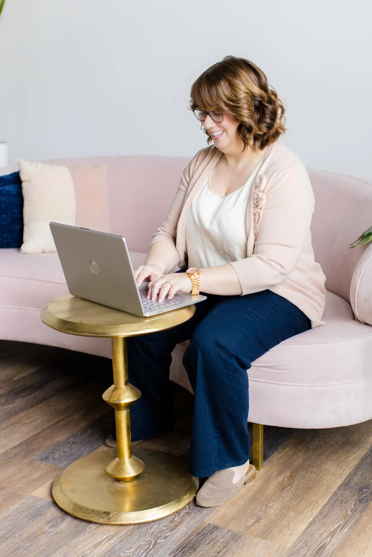 A smiling woman with glasses sitting on a light pink sofa, working on a silver HP laptop placed on a small round gold side table in a living room.