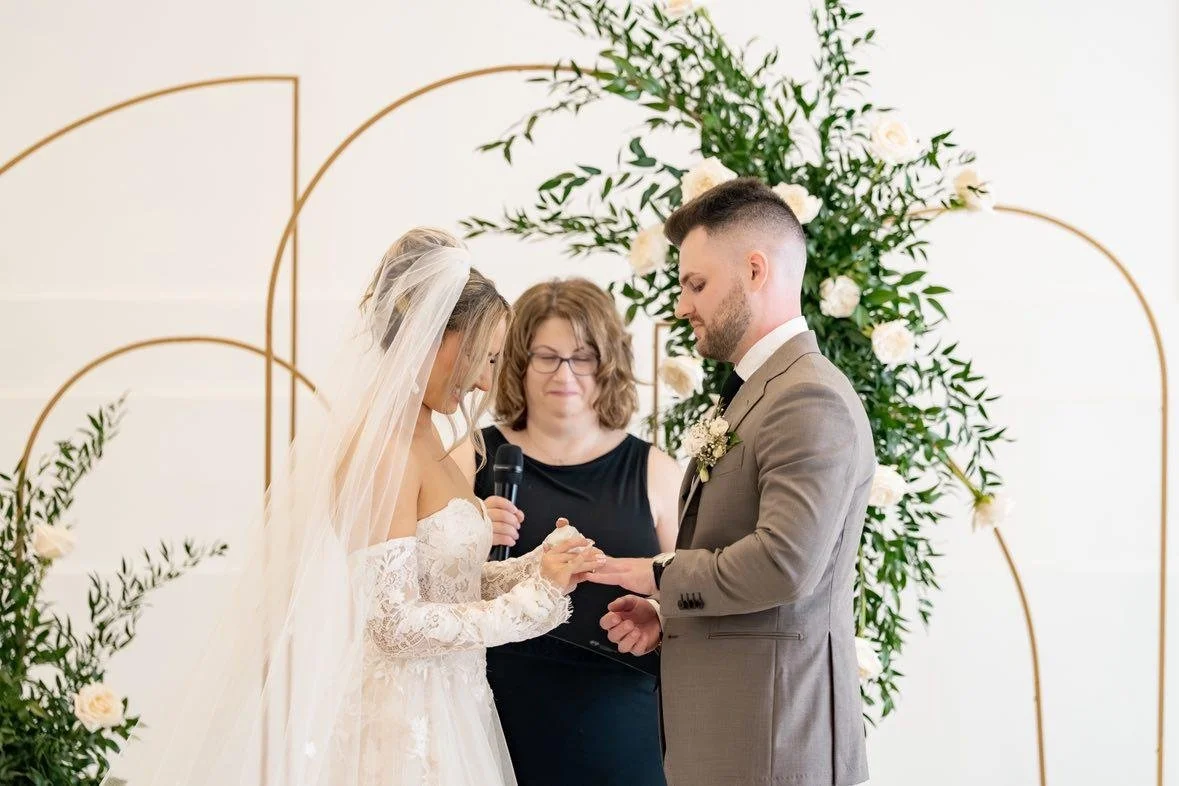 Bride and groom exchanging rings during a wedding ceremony with an officiant standing behind them, floral arch in the background.