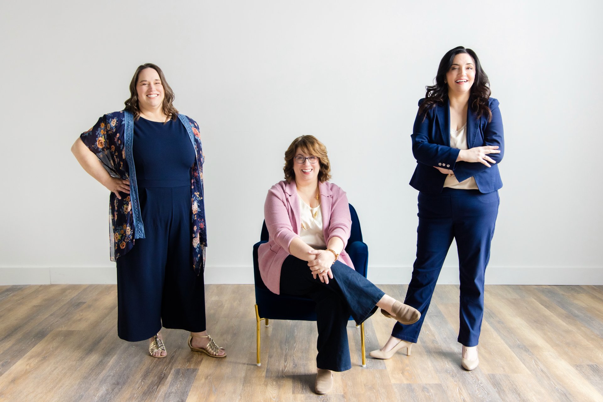 Three women together in a room with a plain white wall; one woman is seated in the middle on a blue chair, and two women are standing on either side of her, all smiling and dressed in business casual attire.