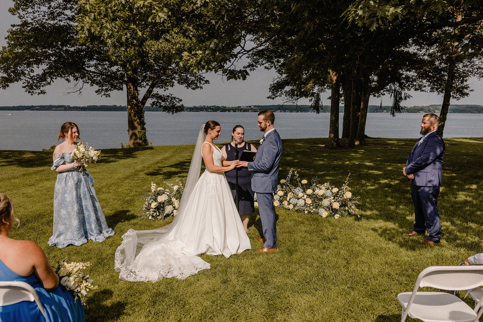 A wedding ceremony taking place outdoors by a lake, with the bride and groom exchanging rings while an officiant conducts the ceremony. The bride is in a white gown with a veil, and the groom is in a blue suit. Bridesmaids and groomsmen stand nearby,