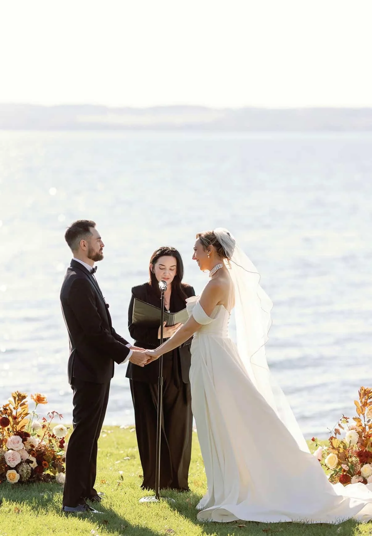 A bride and groom holding hands during a wedding ceremony outdoors near a body of water, with a officiant reading vows, and floral arrangements on the grass.