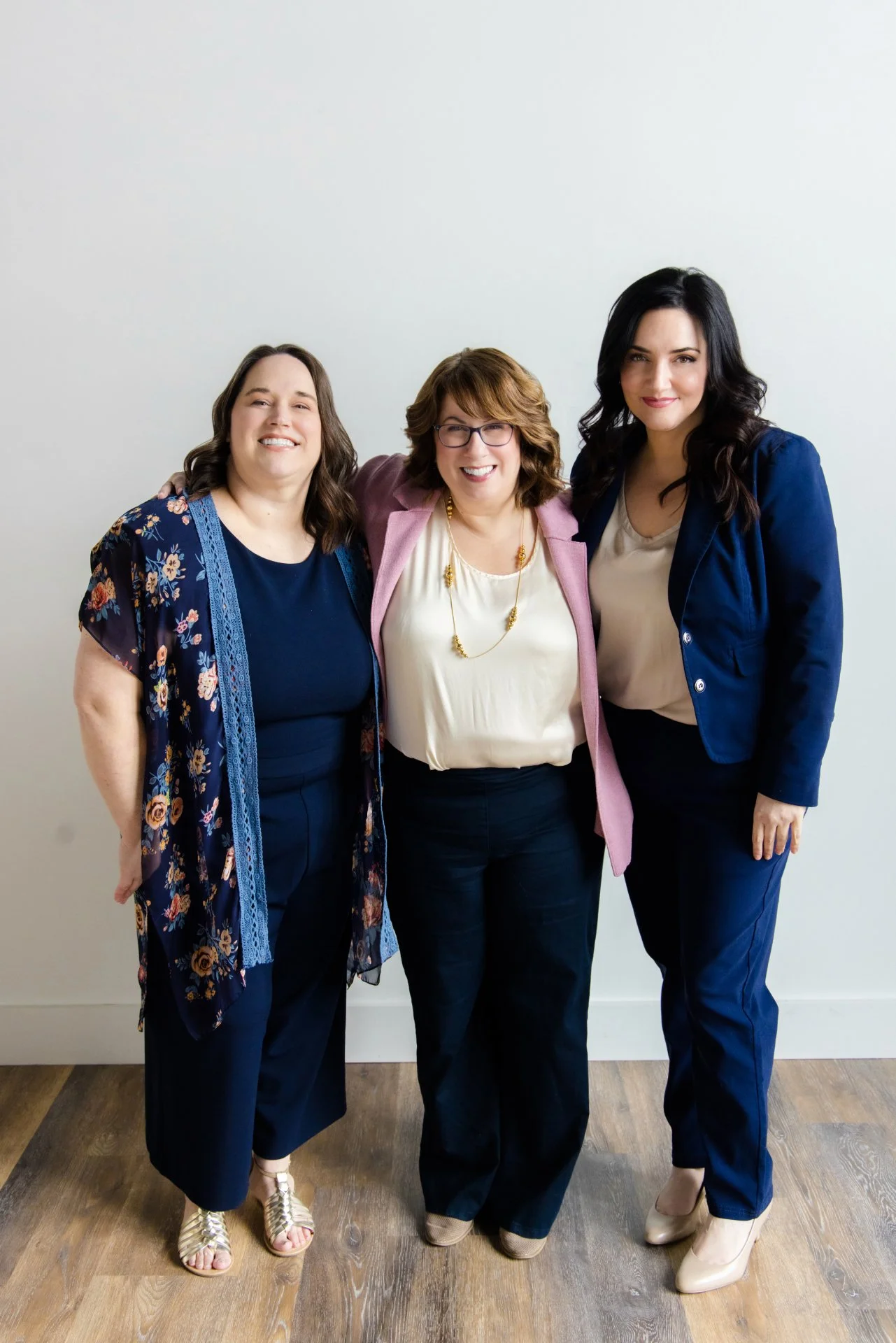 Three women standing together, smiling, in front of a plain wall.