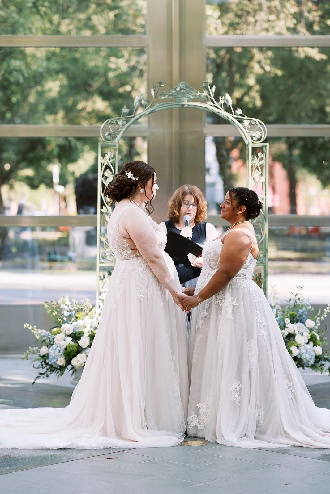 Two women in wedding dresses holding hands during a wedding ceremony, with an officiant behind them reading from a book outdoors, in front of large glass windows and surrounded by floral arrangements.