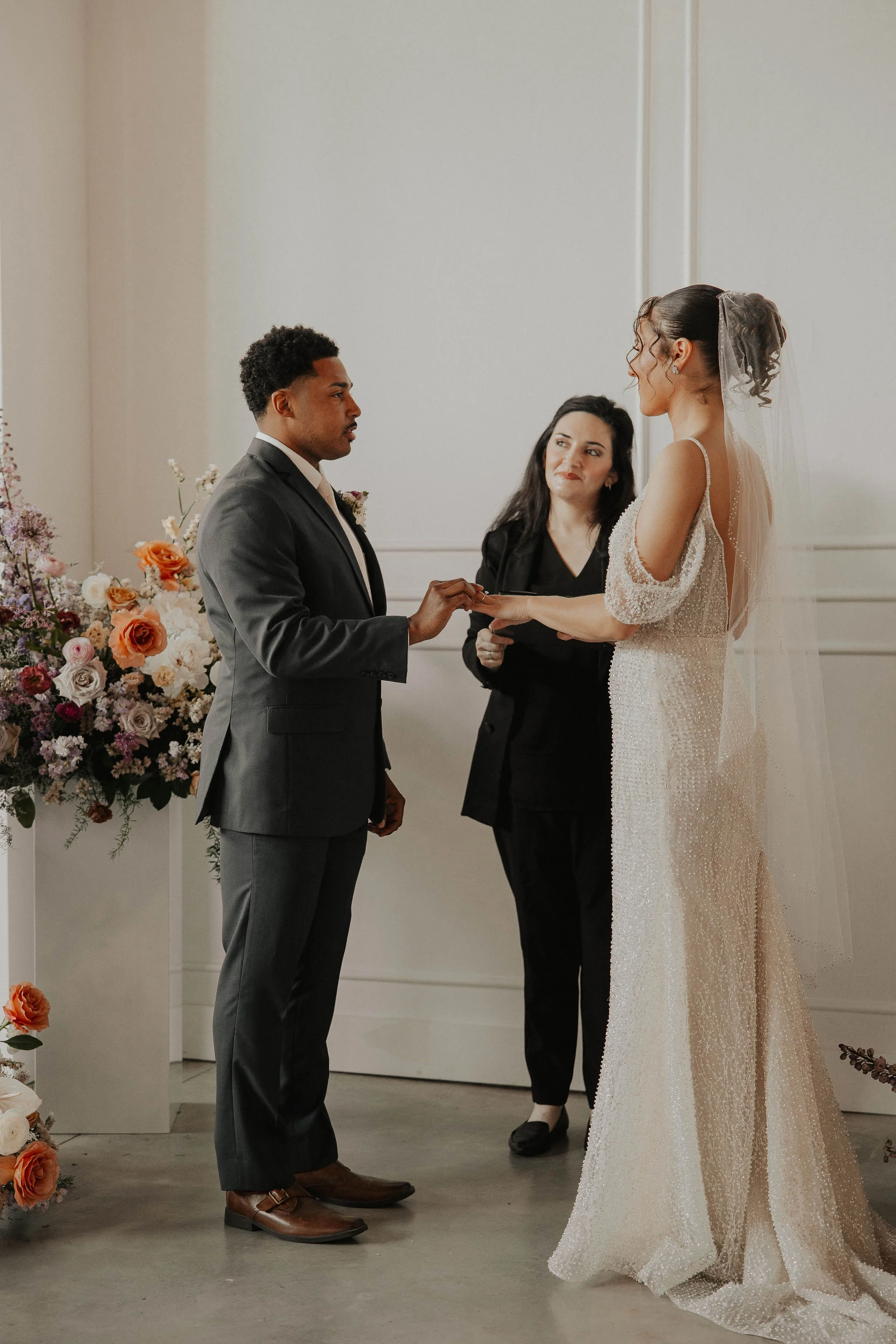 Man in a grey suit with a woman in a wedding dress holding hands in front of a female officiant surrounded by flowers