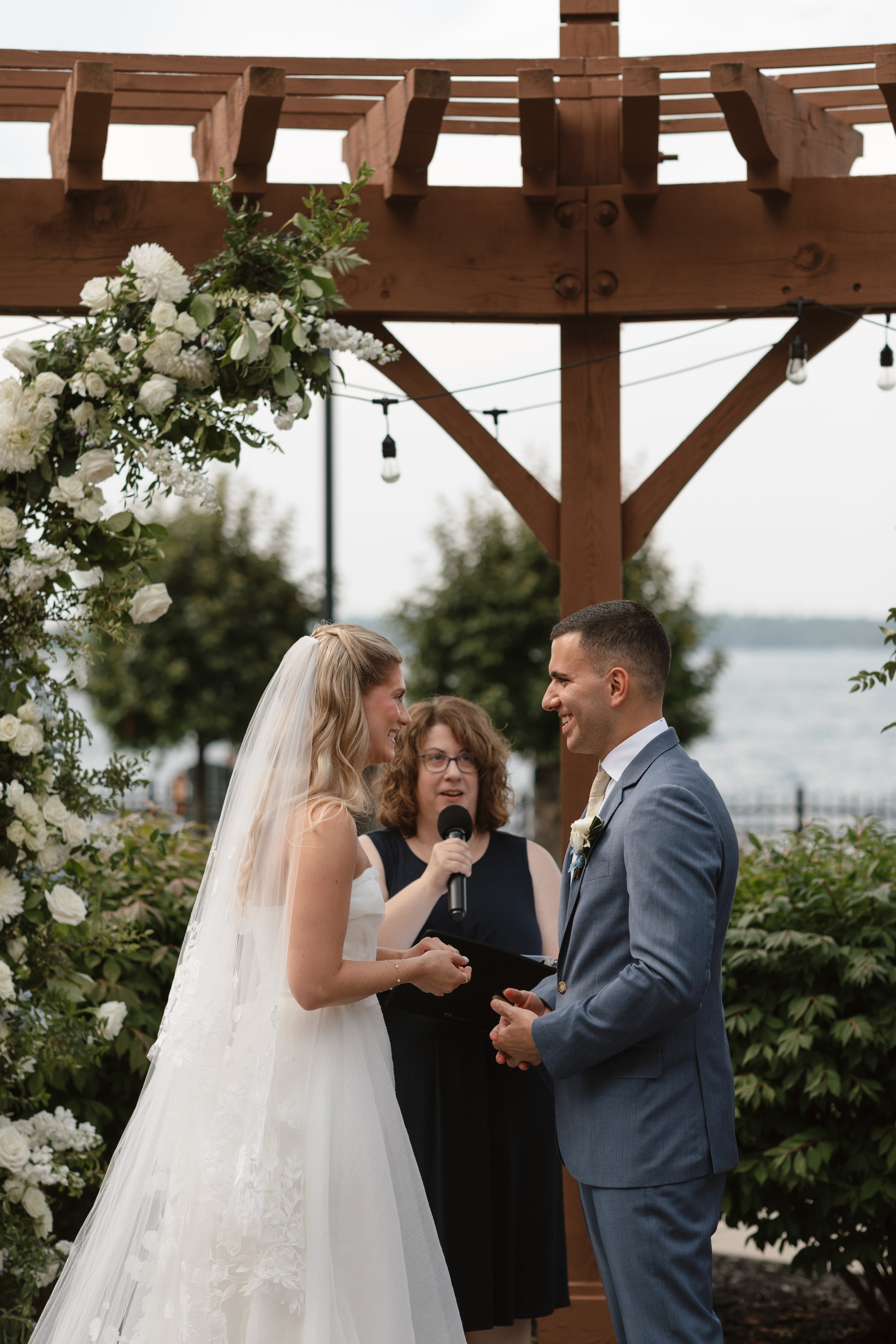 A bride and groom exchanging vows under a wooden wedding arch decorated with white flowers and greenery, outdoors near a body of water, with a officiant holding a microphone.