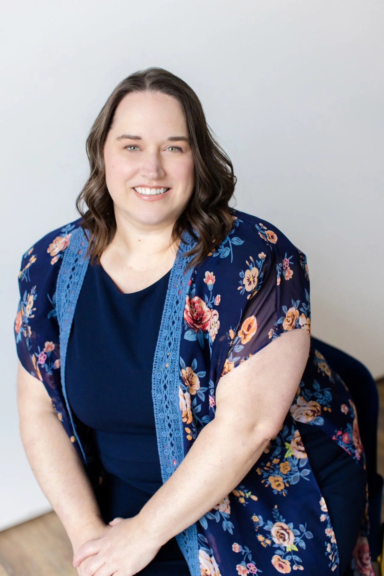 A woman with shoulder-length brown hair, light skin, and blue eyes smiling at the camera. She is wearing a navy blue dress and a floral kimono with blue lace trim, sitting on a blue chair against a plain light background.