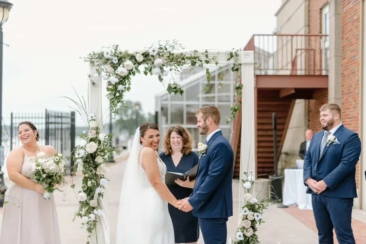 A bride smiling at her guests while holding hands with her groom and officiant in navy blue holding a binder smiling